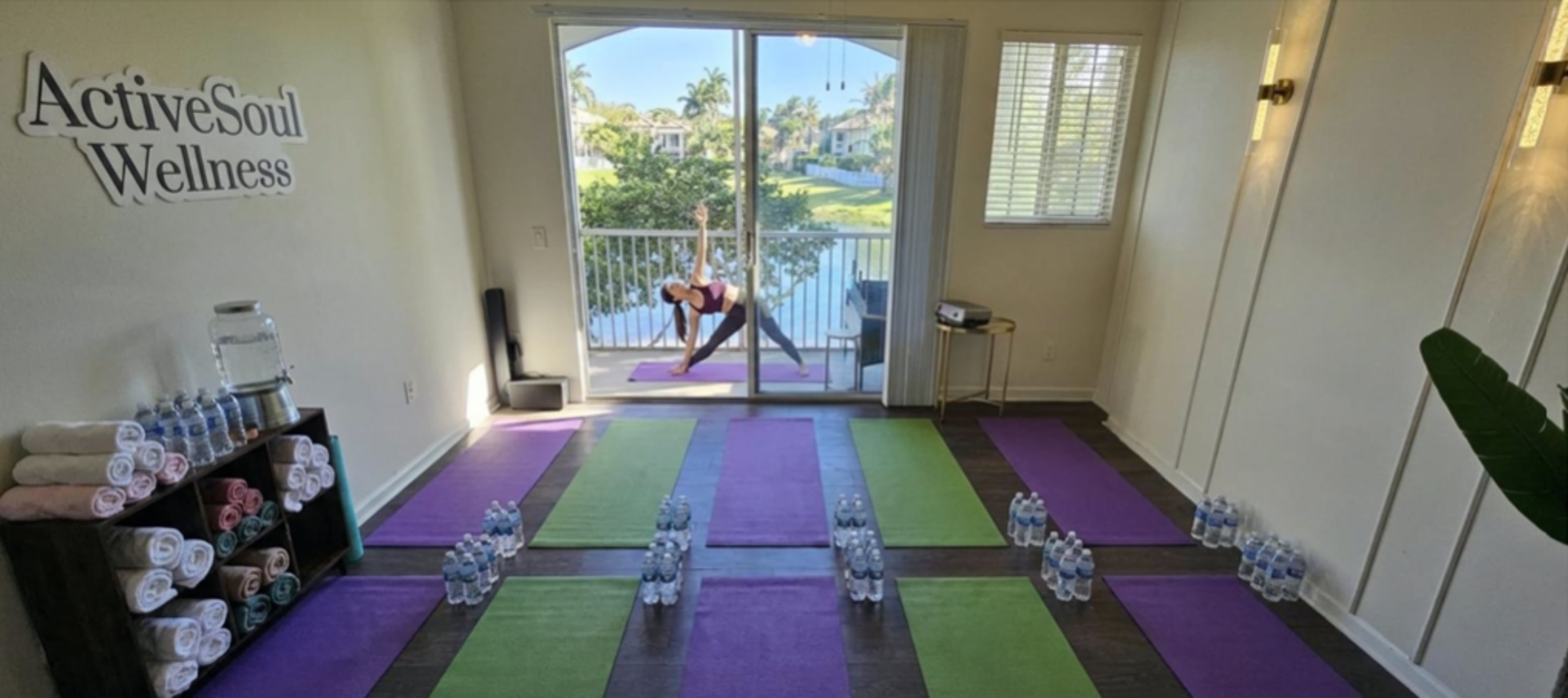 The image shows a yoga studio with colorful mats arranged on the floor and a person practicing yoga near a large window that overlooks a body of water.