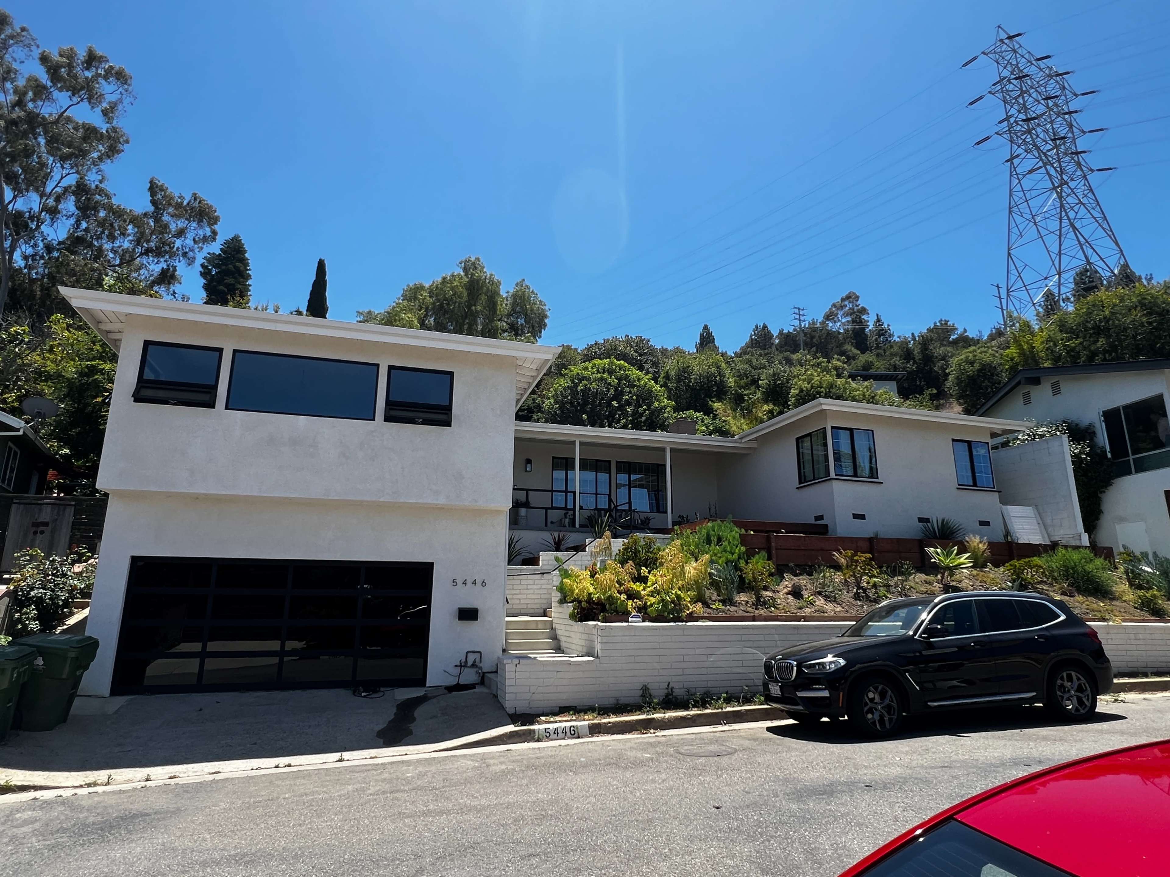 The image shows a modern white house with a black garage door, surrounded by greenery, located on a residential street under a clear blue sky.