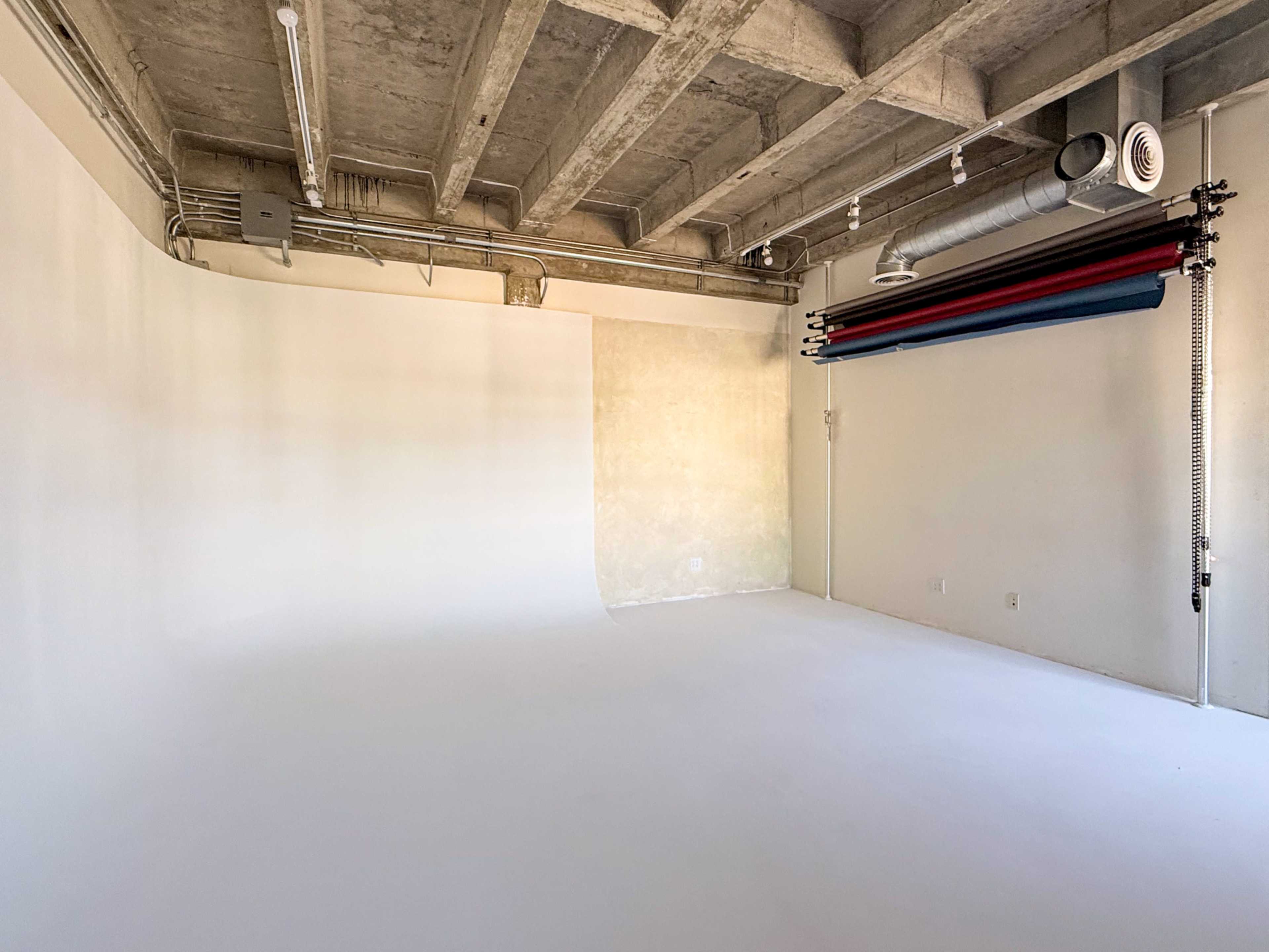 The image shows a minimalist photography studio with a white seamless backdrop, concrete walls, and a row of colored backdrop rolls at the top.