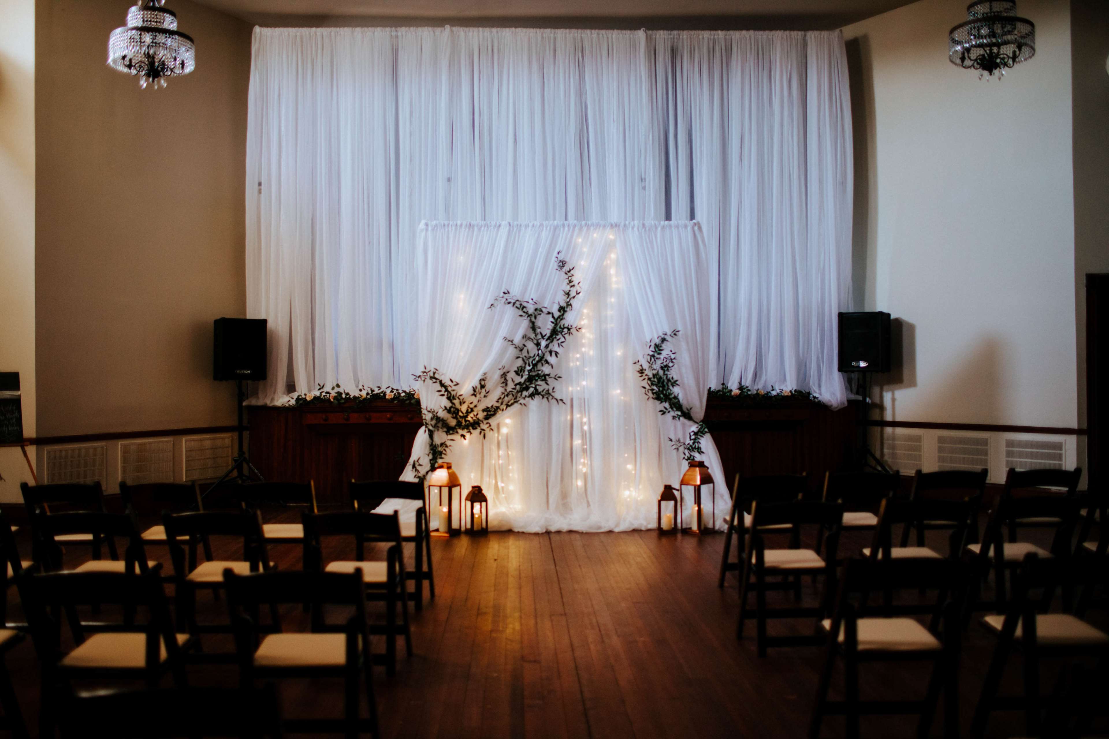 A decorated ceremony space features a white backdrop adorned with fairy lights, flanked by plants, and rows of wooden chairs facing the setup.