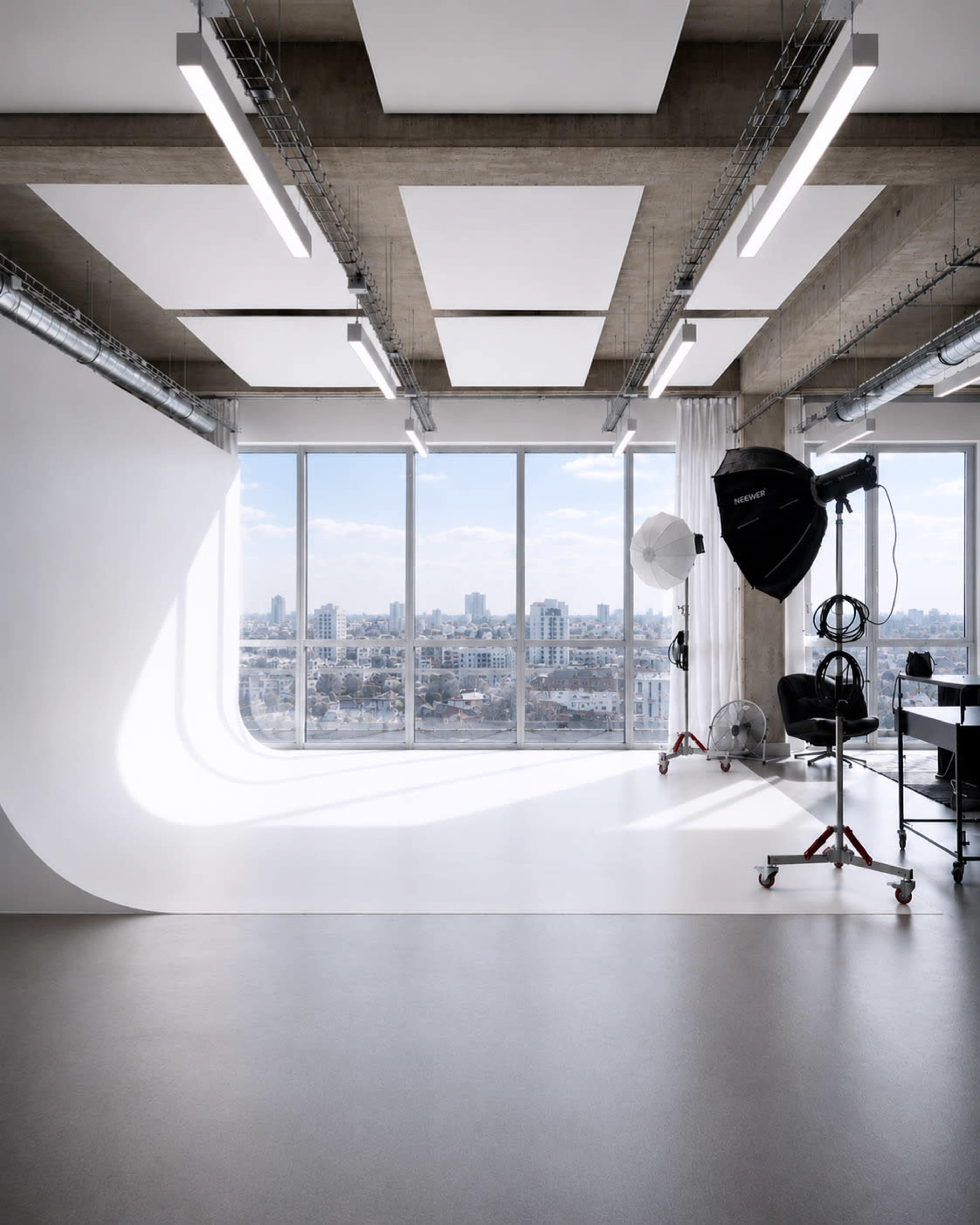 The image shows a modern photography studio with large windows providing a view of a city skyline, featuring a seamless white backdrop and studio lighting equipment.