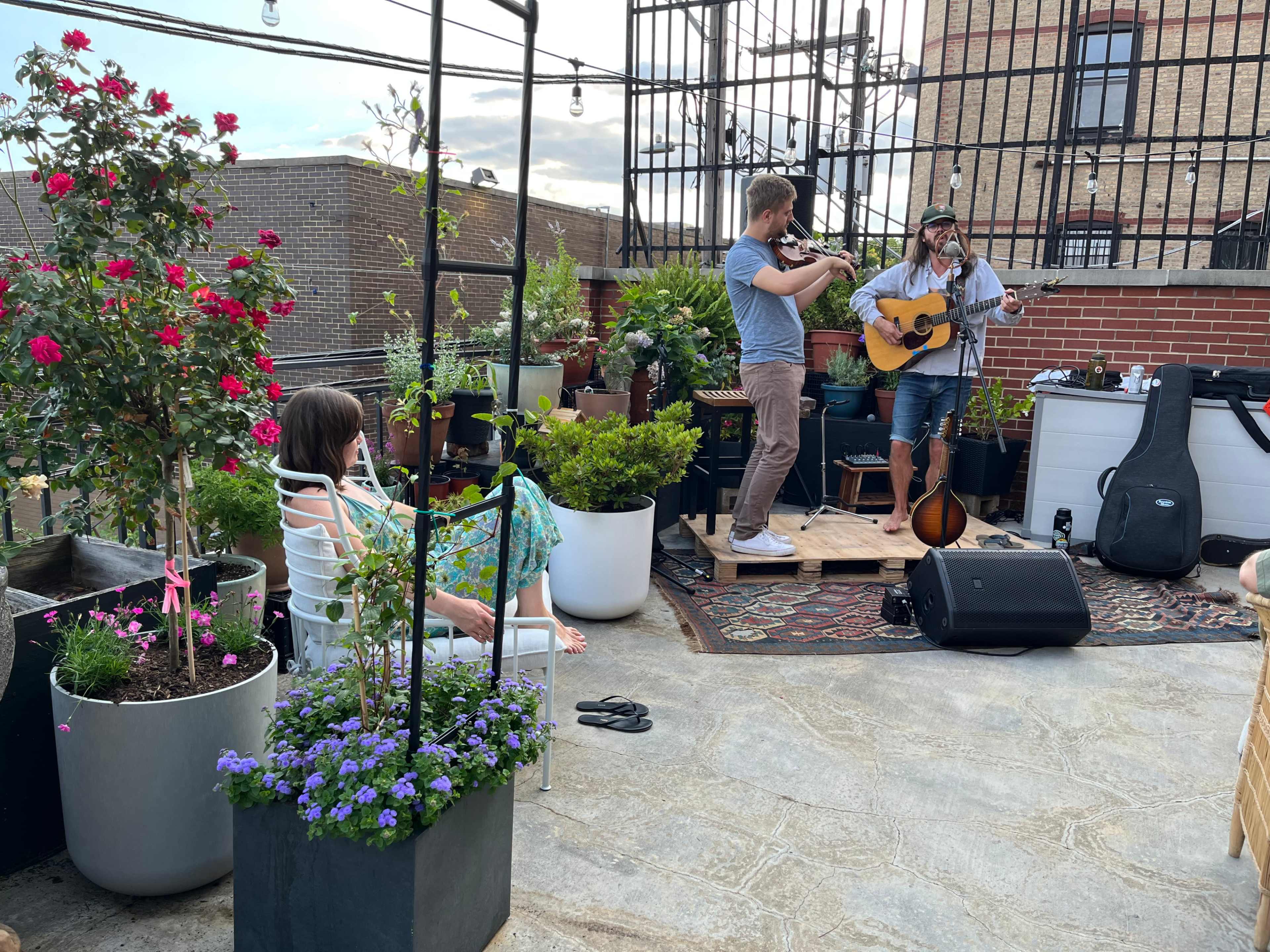 Two musicians perform on a rooftop surrounded by plants, while a woman relaxes in a chair nearby.