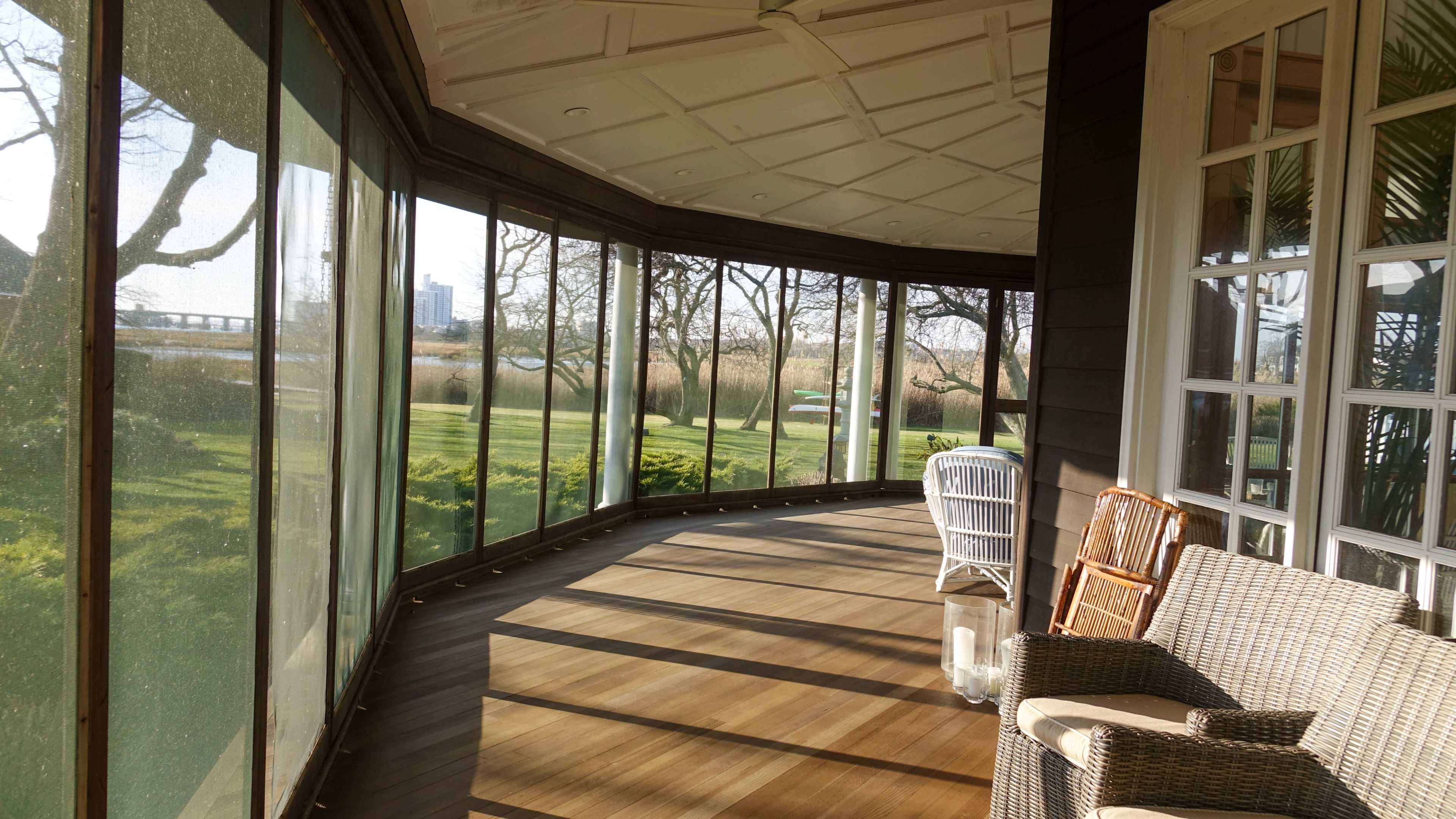 A sunlit enclosed porch with wooden flooring, featuring a white wicker chair and a woven chair, overlooking a green lawn and trees.