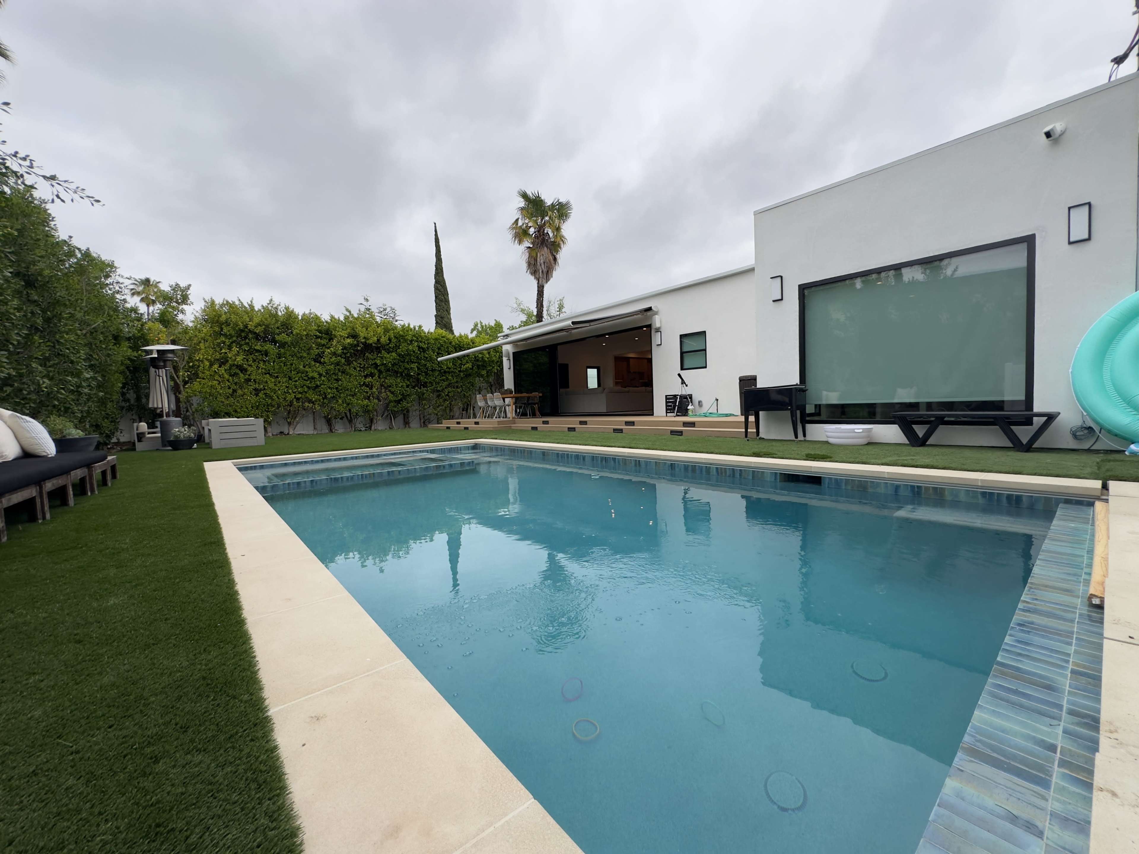 The image shows a swimming pool surrounded by artificial grass, with a modern house and patio in the background under a cloudy sky.
