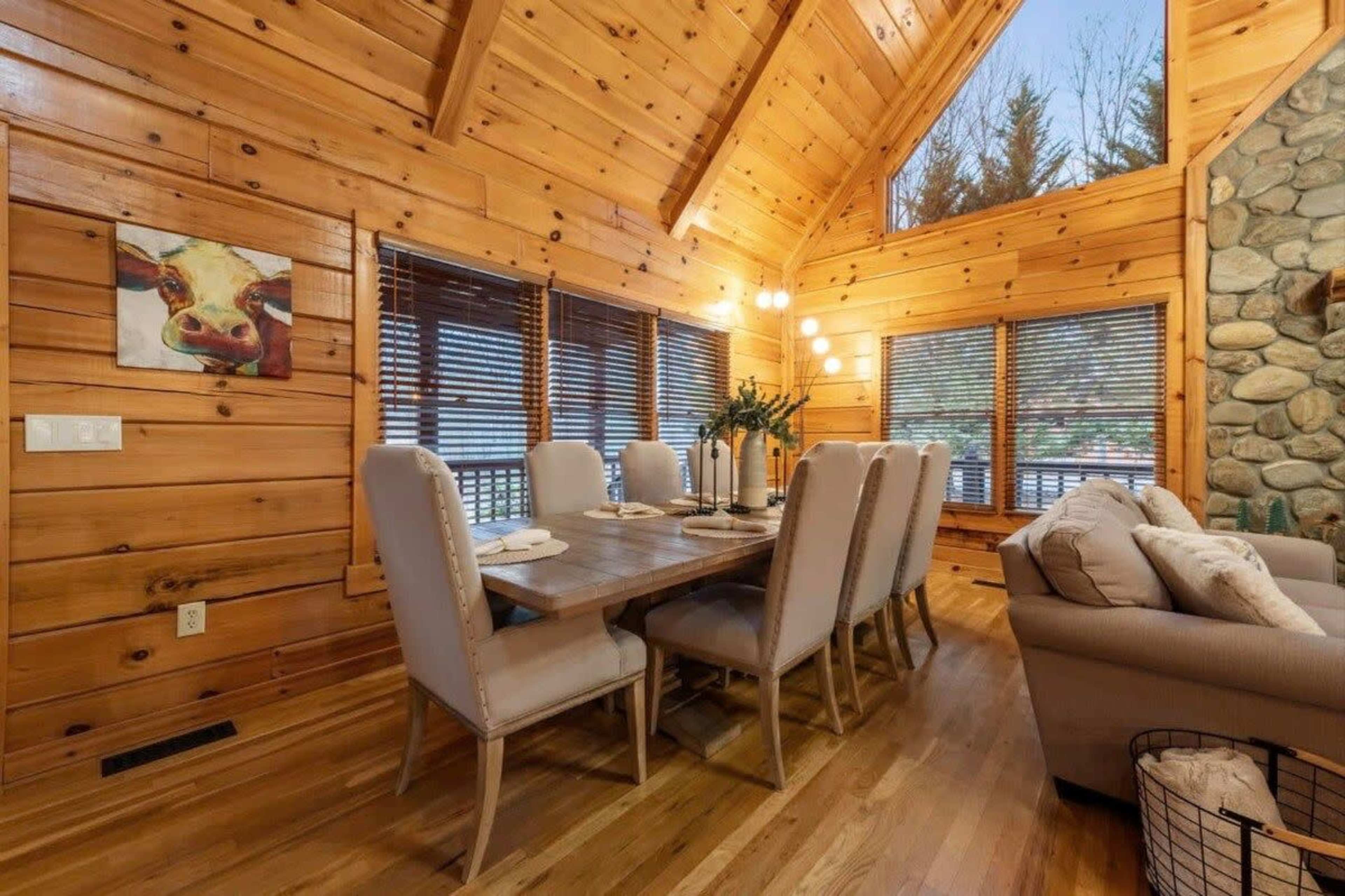 The interior of a wooden cabin features a dining table set for eight, surrounded by upholstered chairs, with large windows allowing natural light in.