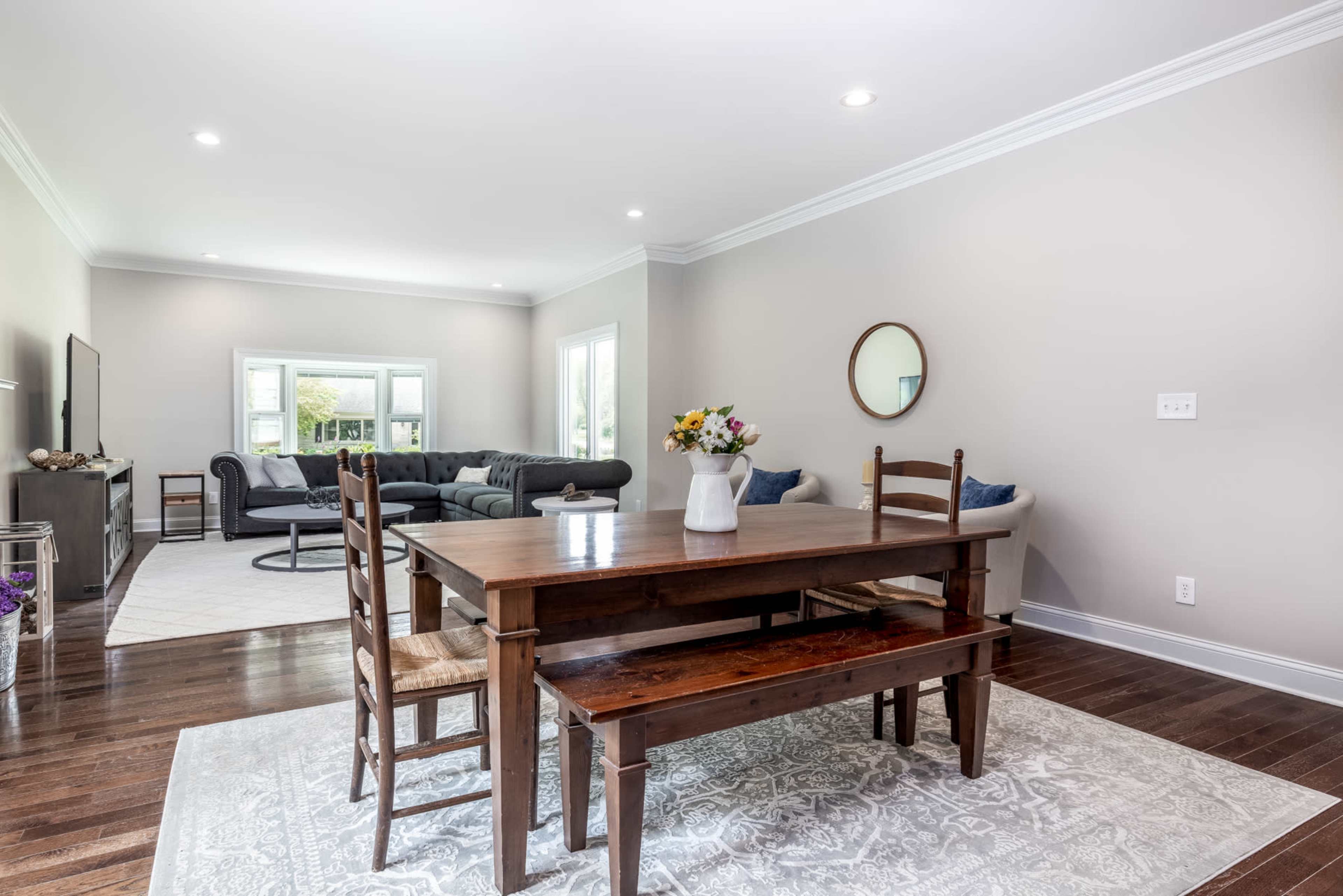 A wooden dining table with chairs sits on a patterned rug in a bright, spacious living area featuring a gray sofa and large windows.