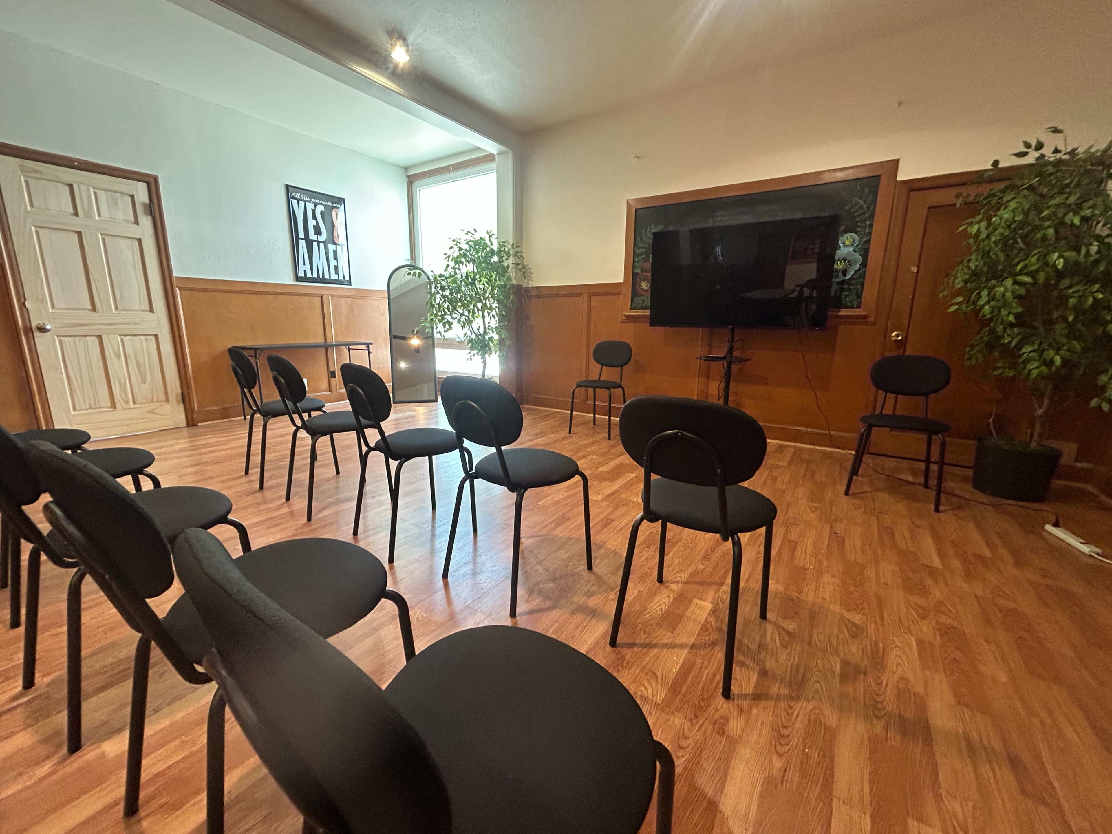 The image shows a room with wooden flooring, featuring several black chairs arranged in a circle, a large television screen, and two plants in the corners.