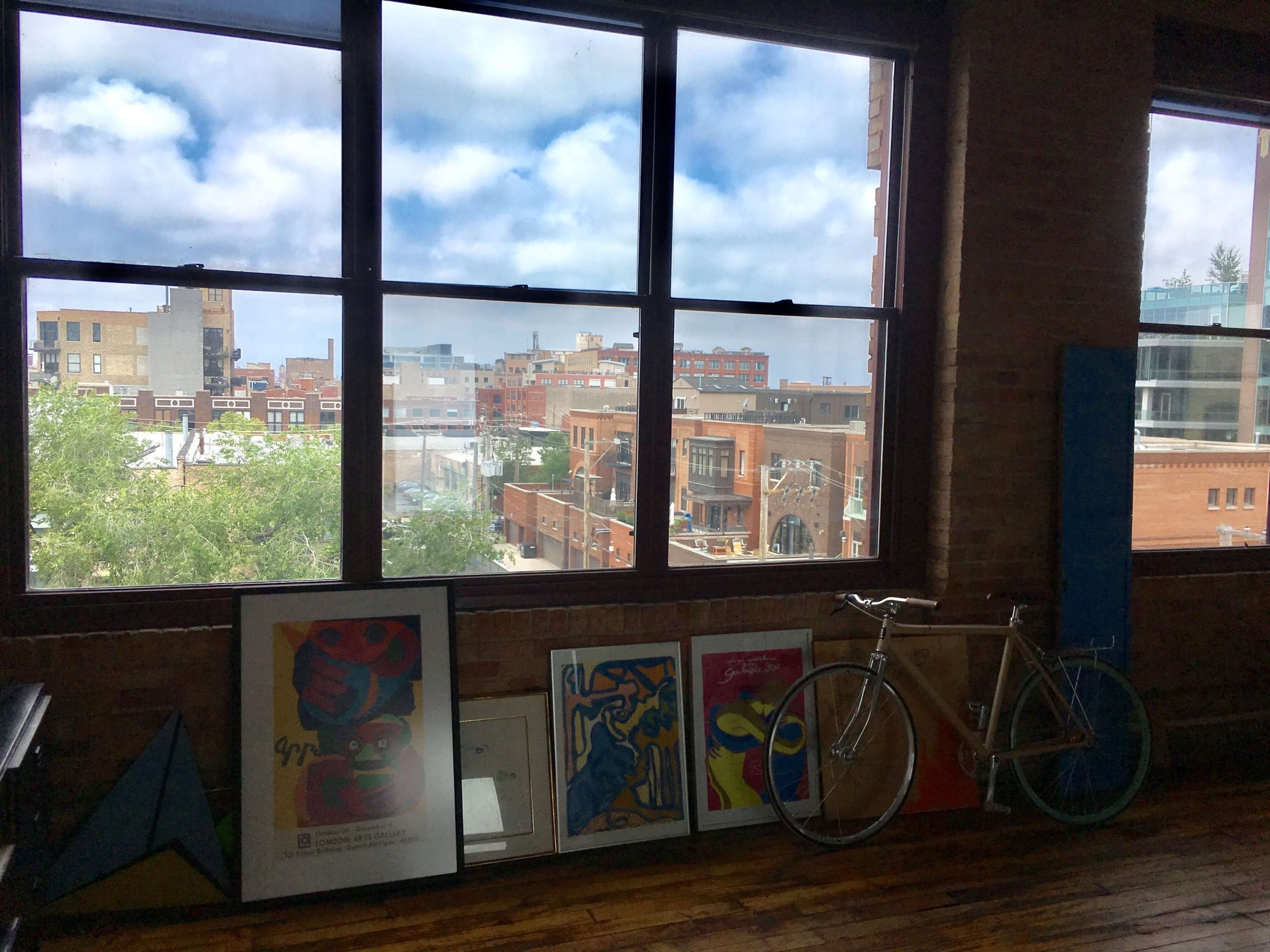 A room with large windows overlooking a cityscape, featuring several framed artworks and a bicycle leaning against the wall.