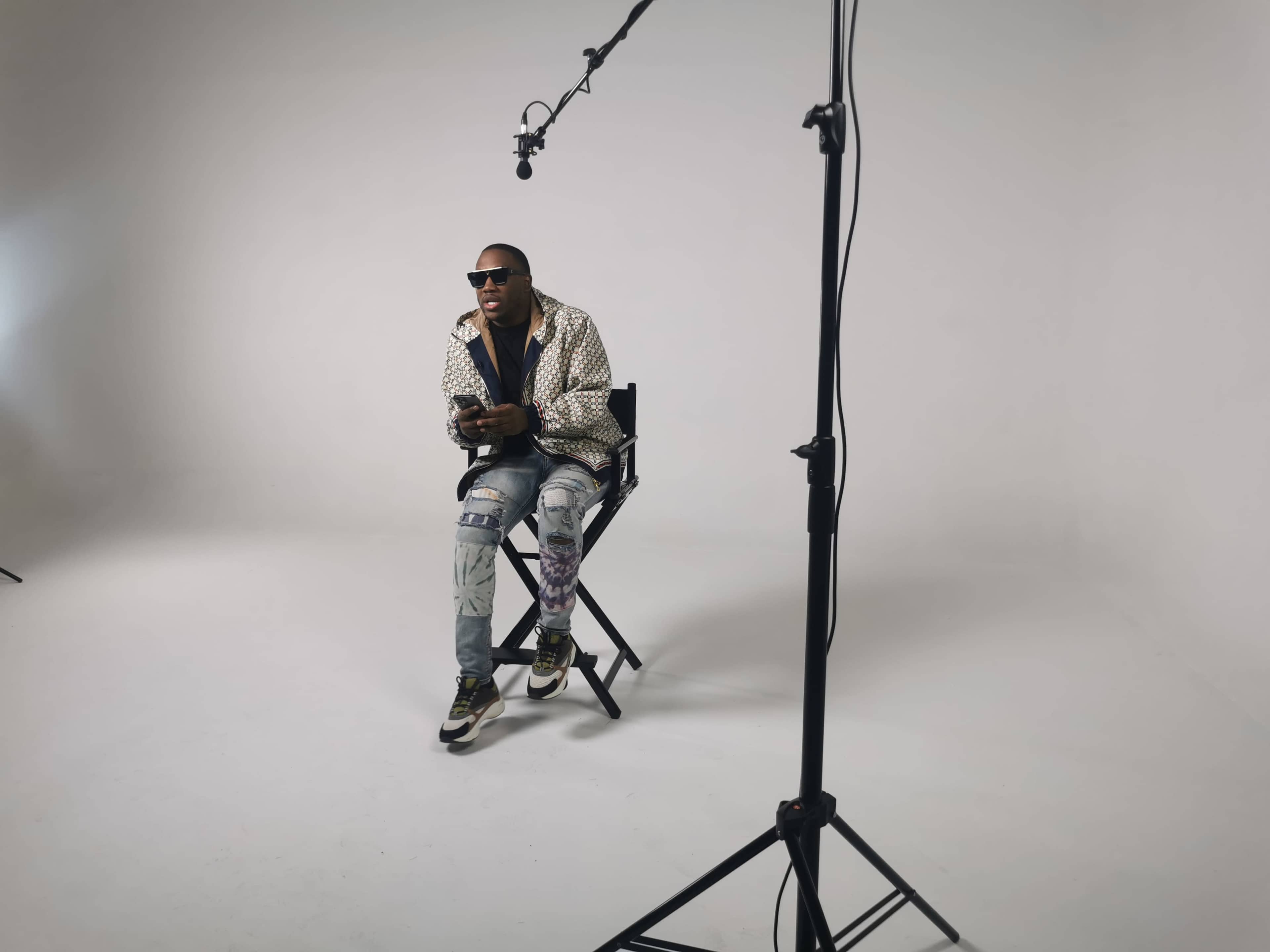 A person sits on a chair in a studio setting, with a microphone on a stand positioned above them and a plain backdrop.
