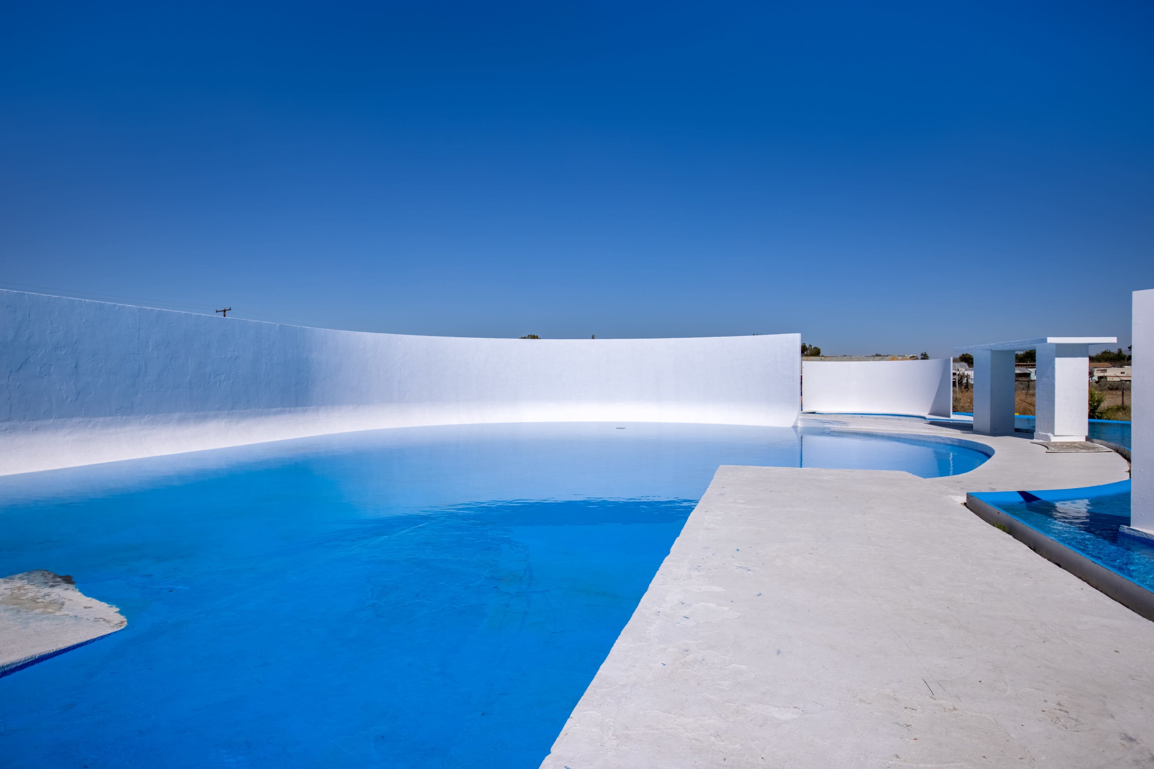 A large, modern swimming pool with bright blue water and white surrounding walls is visible under a clear blue sky.