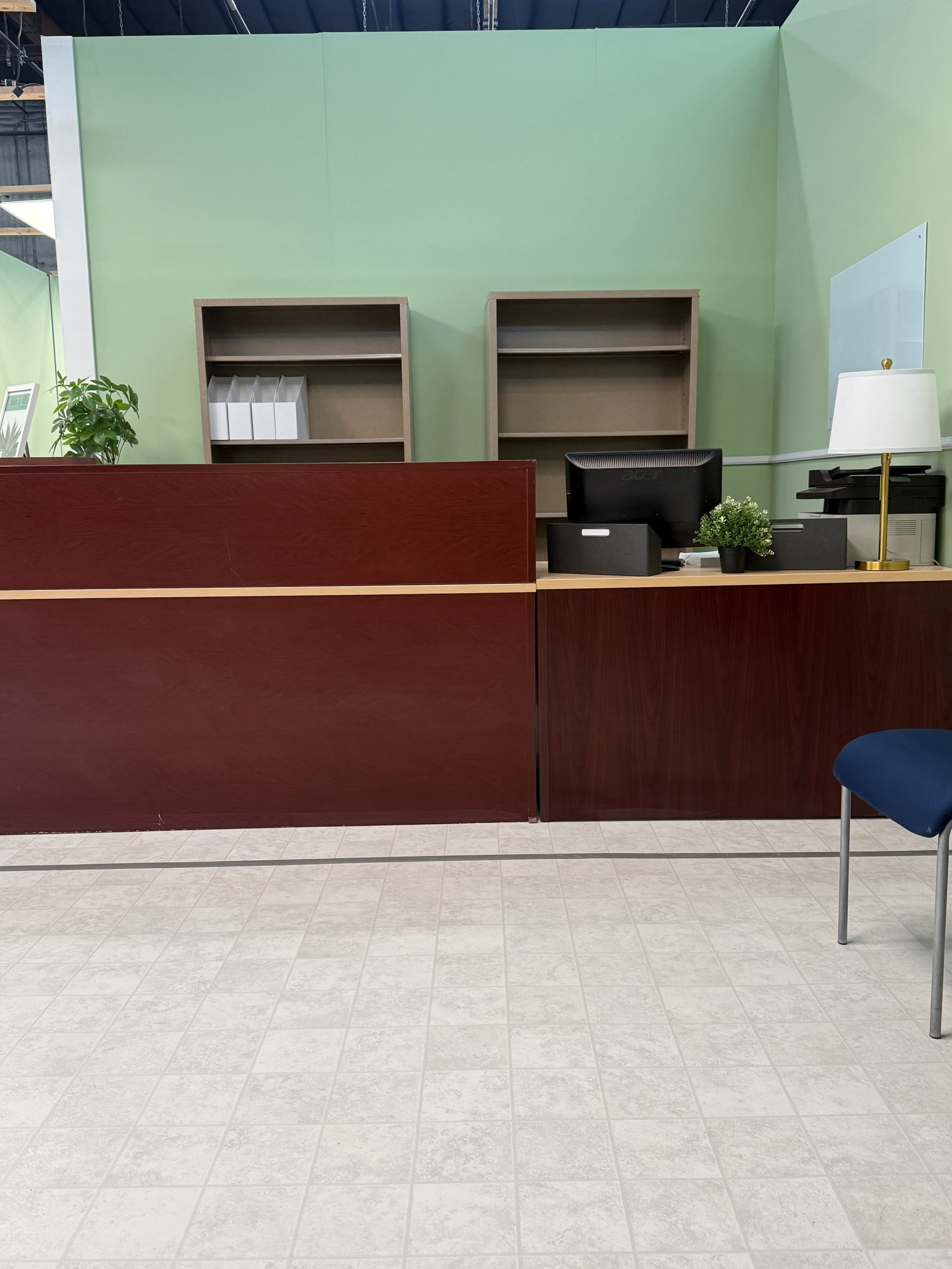 The image shows a reception area with a wooden front desk, a computer monitor, shelves in the background, a lamp, and a chair nearby.