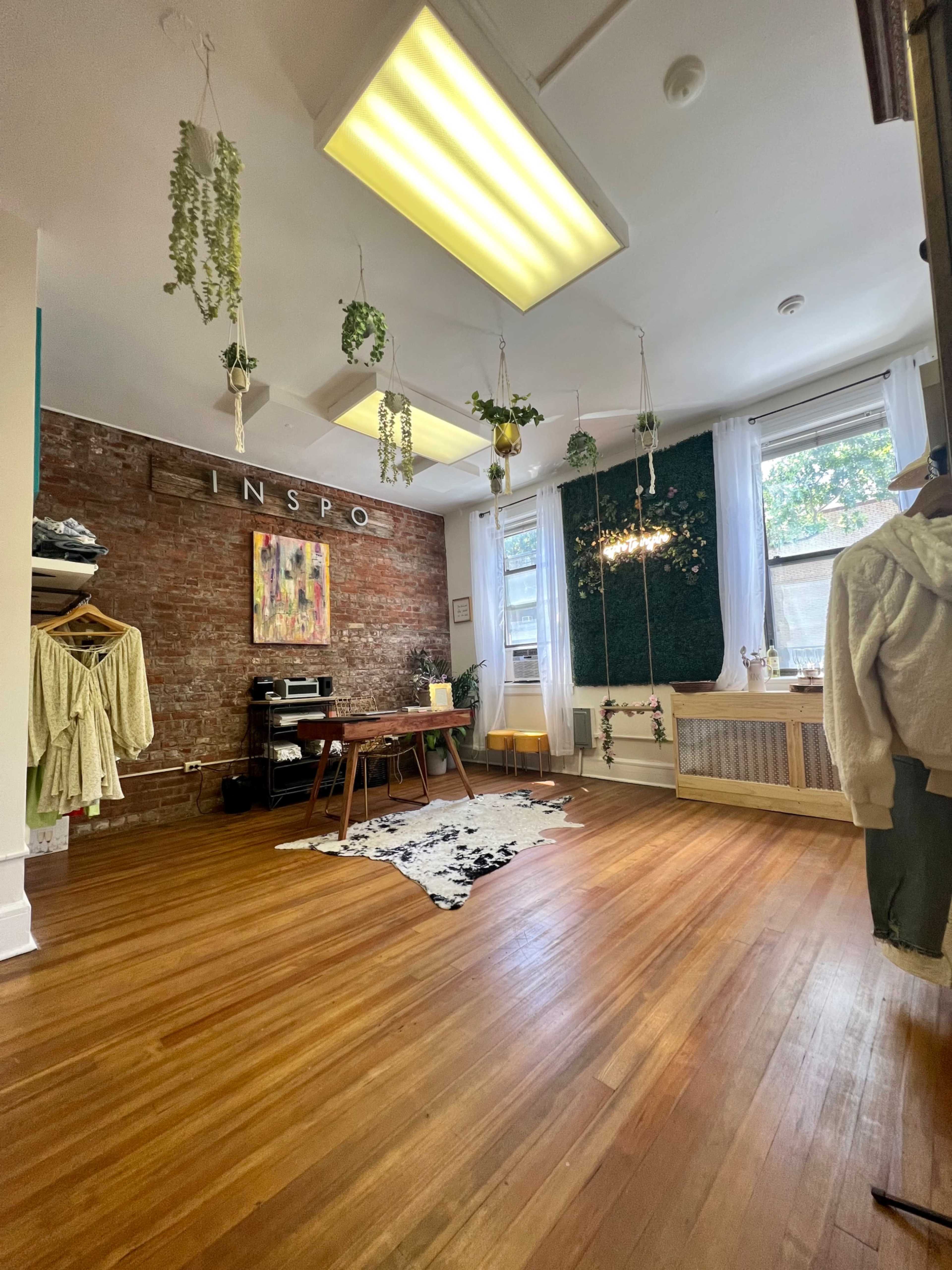 A spacious room with hardwood flooring, featuring a brick wall, hanging plants, and a mix of furniture including a table and clothing racks.