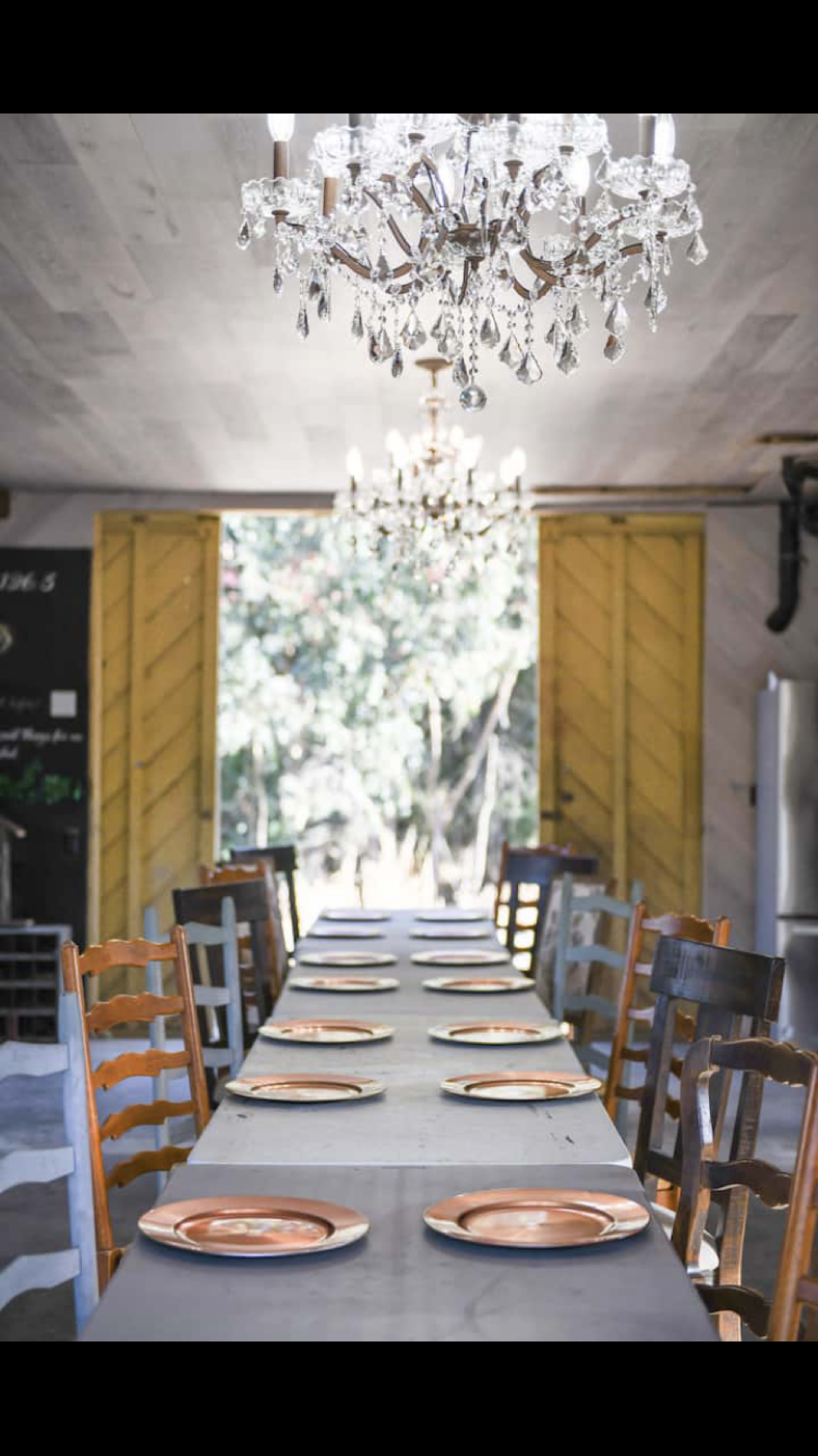 A long dining table is set with plates, flanked by wooden chairs, beneath two chandeliers and a pair of open yellow doors leading to an outdoor view.