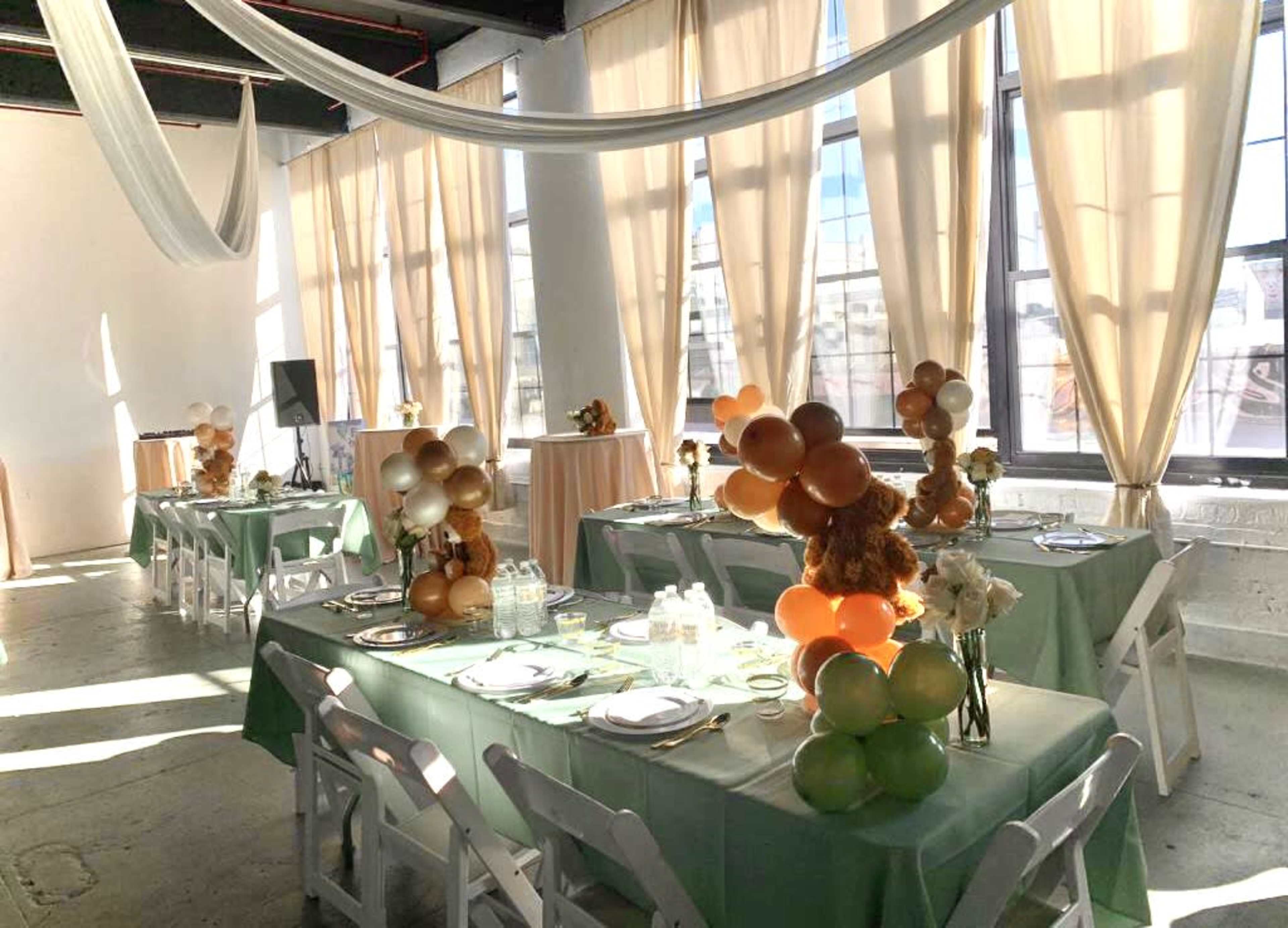 A decorated event space with tables set for a gathering, featuring balloons and floral arrangements under flowing drapes.