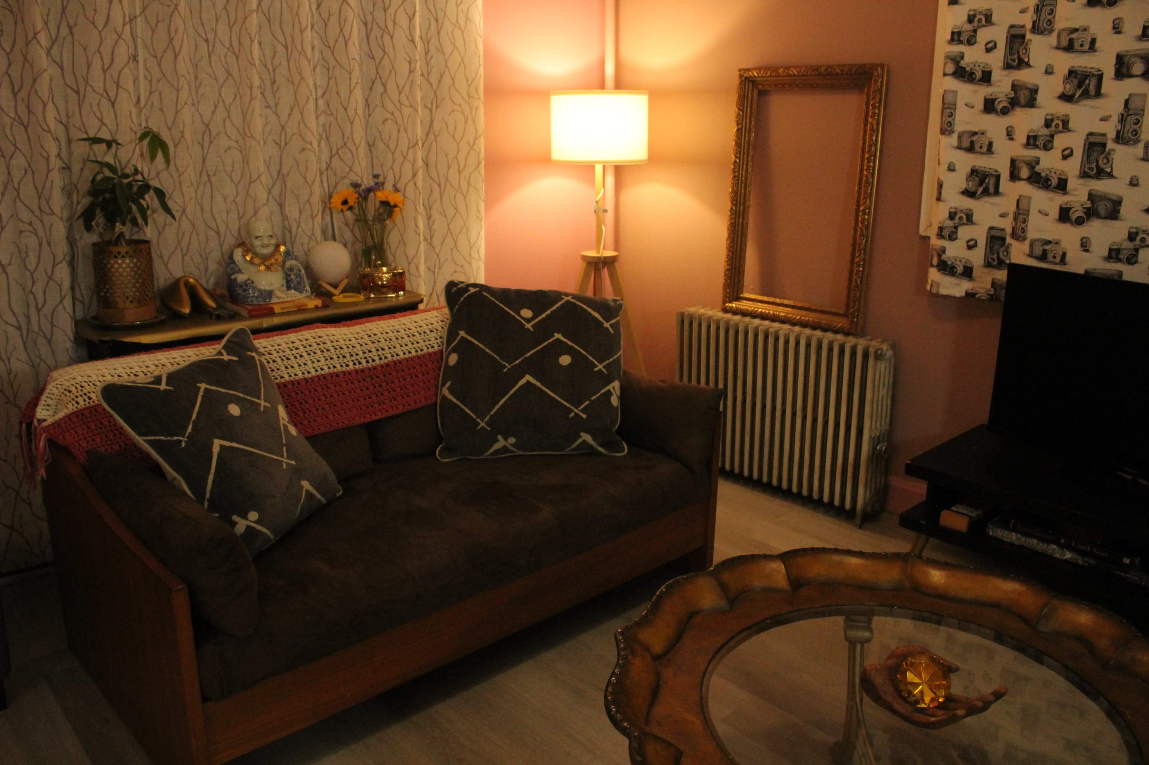 The image shows a cozy living room with a brown couch adorned with decorative pillows, a lamp providing warm light, and various decorative elements, including a framed mirror and a coffee table.