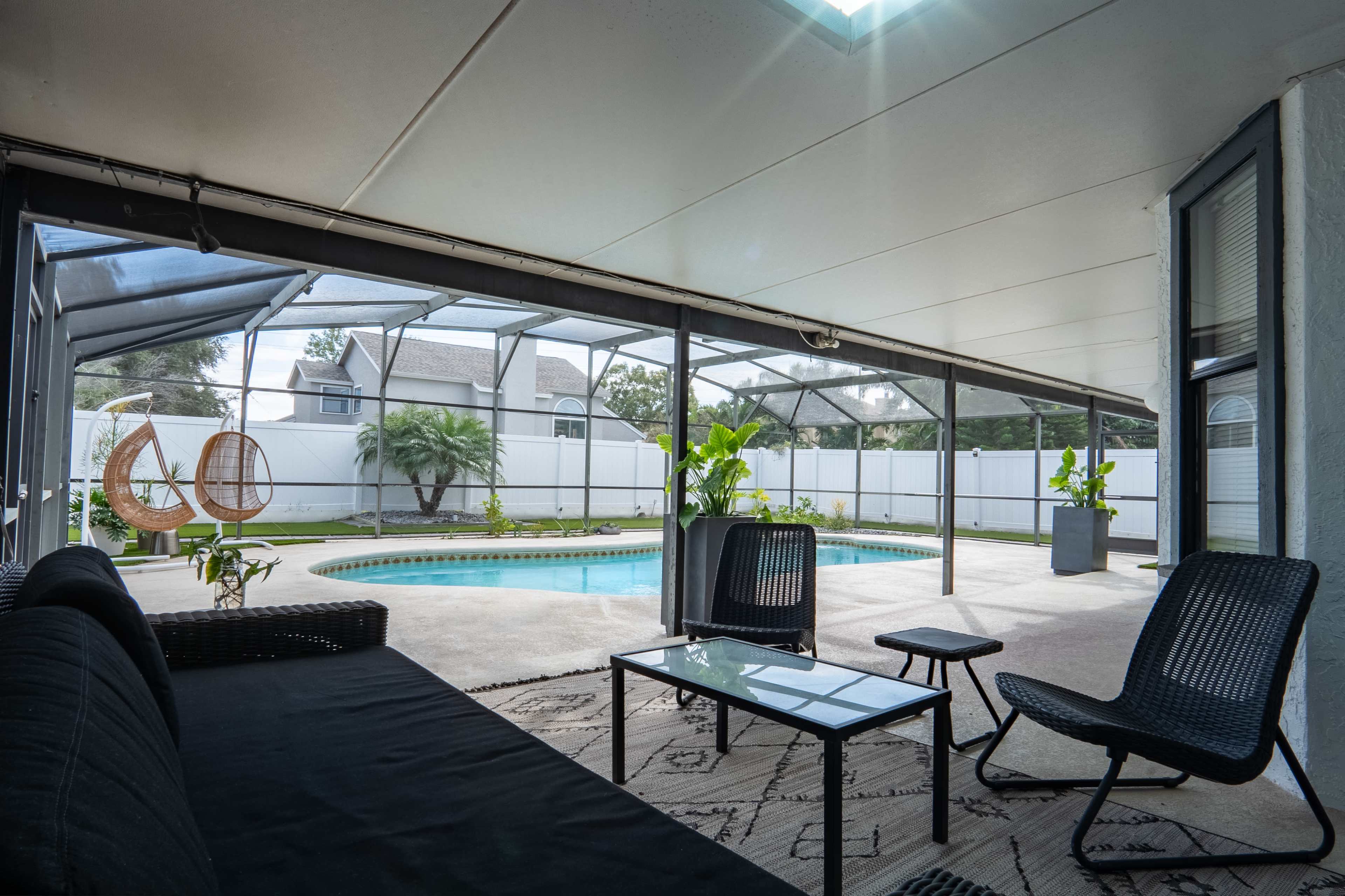 The image shows a covered patio area with a view of a swimming pool surrounded by greenery and plants.