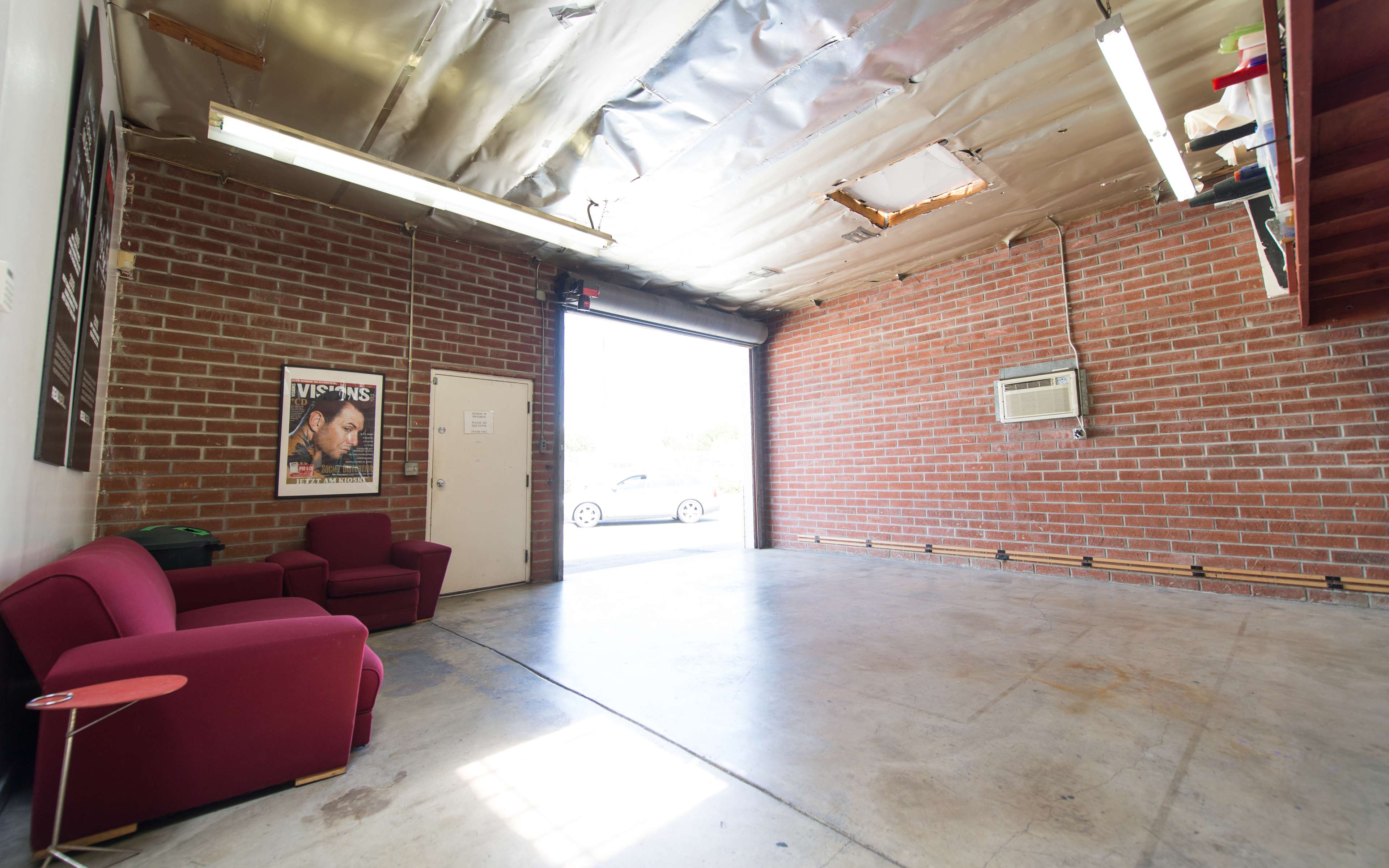 The image shows an empty garage with exposed brick walls, a ceiling with metal tiles, two red chairs, and a door leading outside.