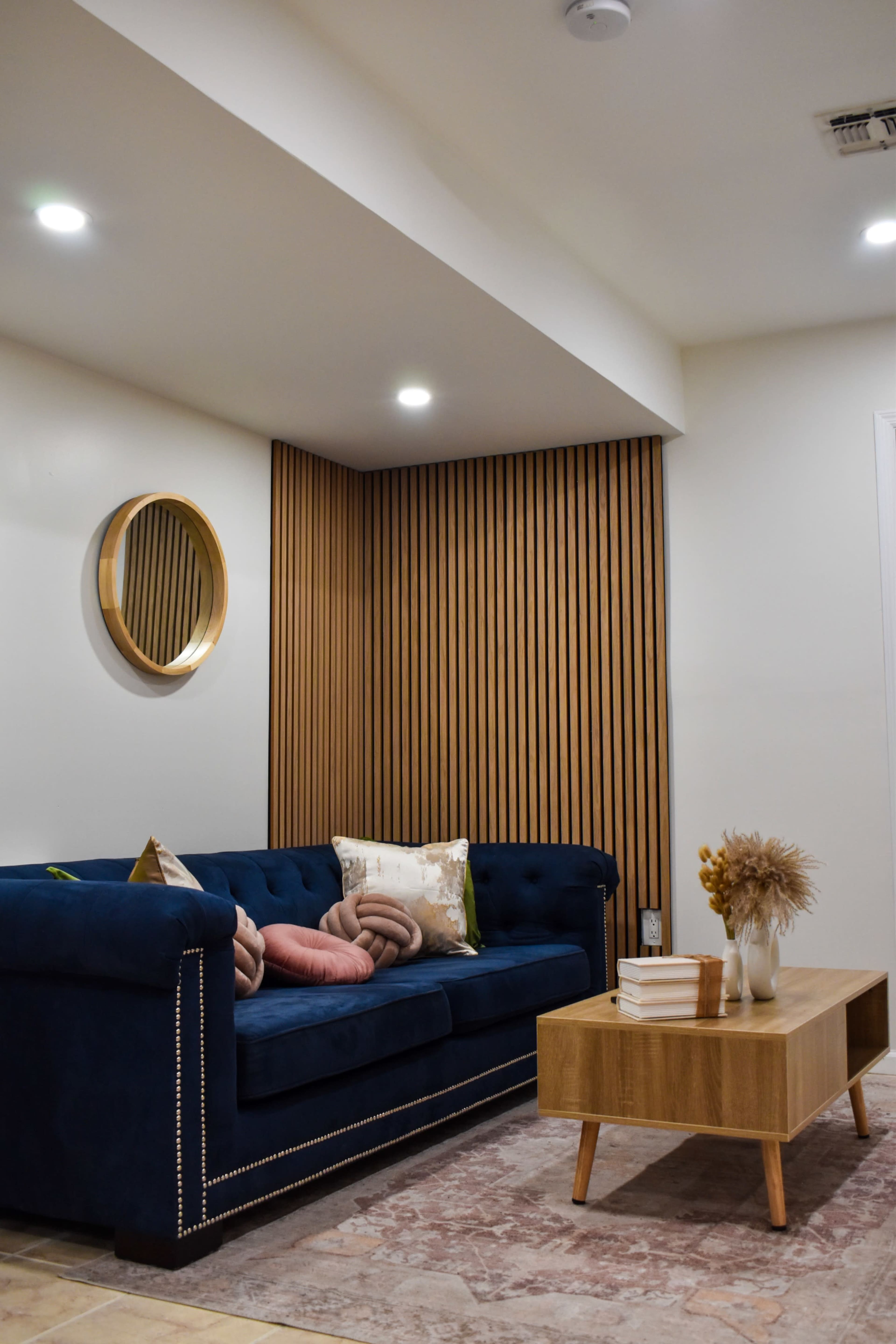 A modern living room features a blue velvet sofa with decorative pillows, a wooden coffee table, and a round mirror above a wooden accent wall.