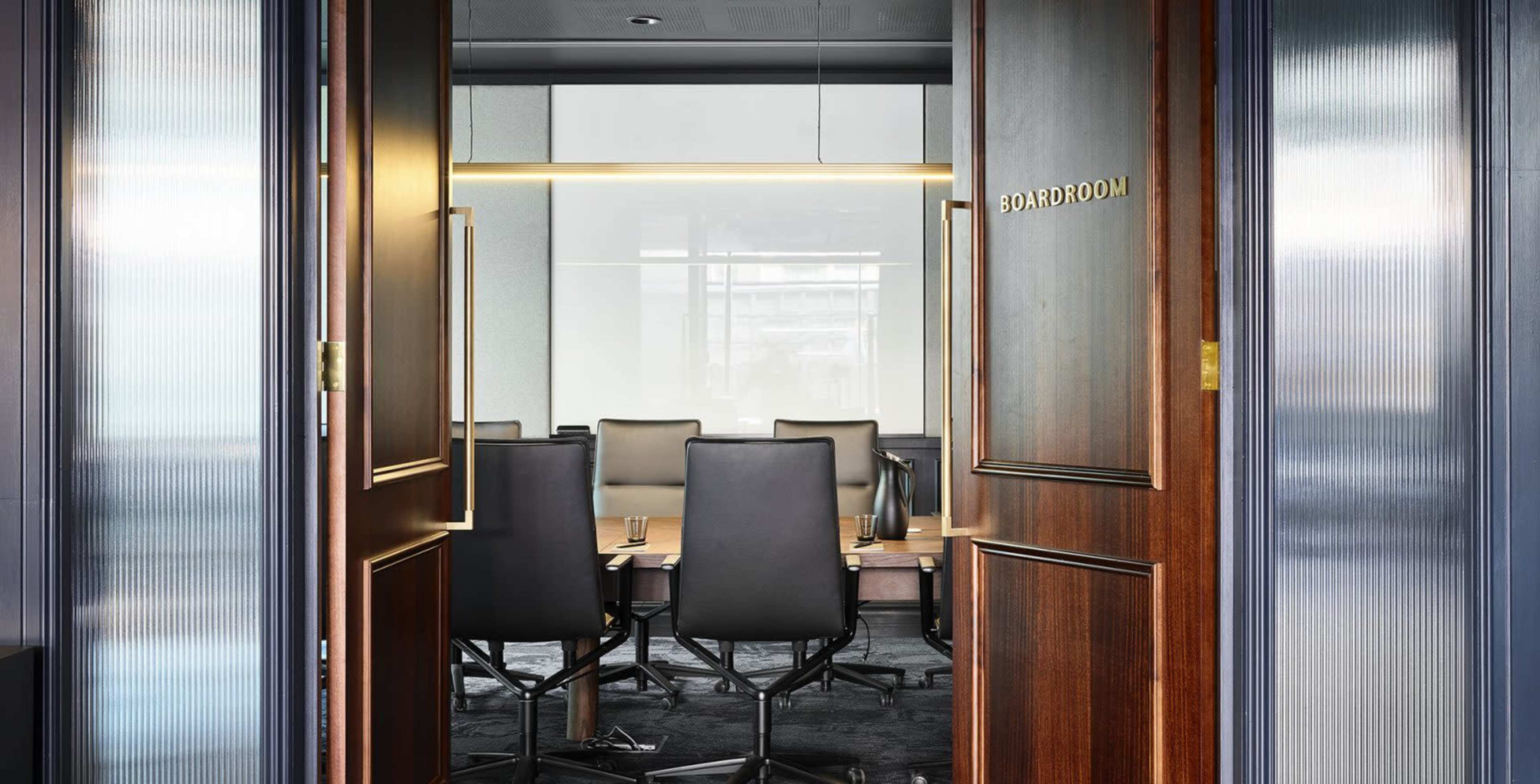 A modern boardroom with a long table and ergonomic chairs, viewed through open wooden double doors.
