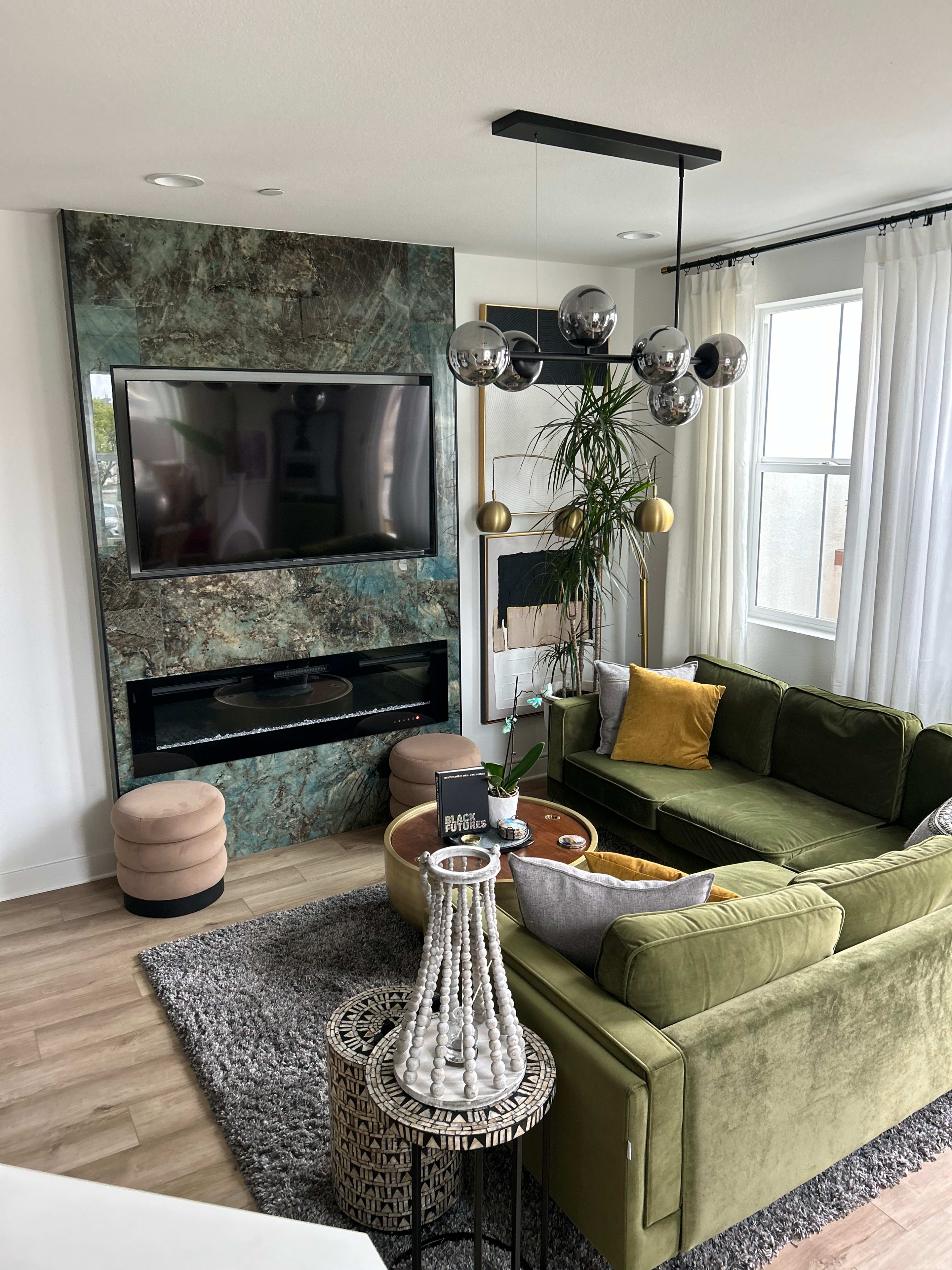 The living room features a green sectional sofa, a large TV mounted on a marbled wall, and a modern light fixture above a coffee table surrounded by stools.