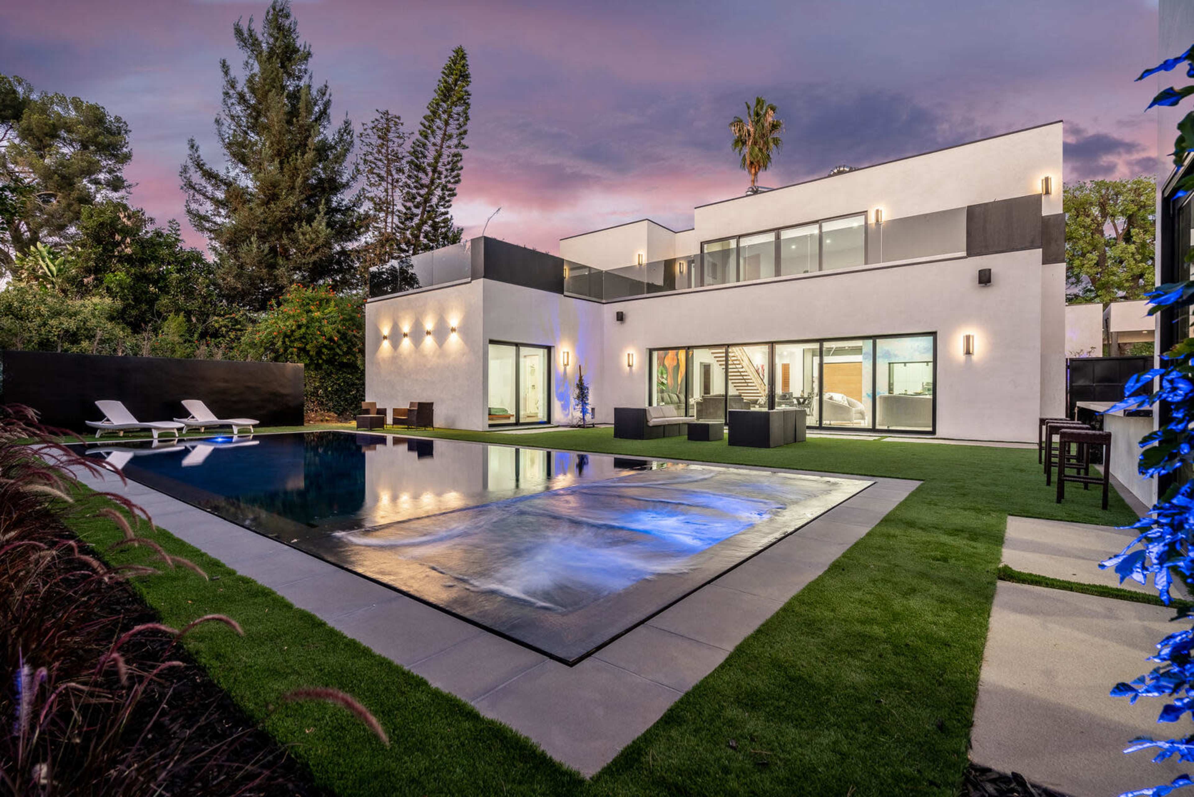 A modern two-story house with large windows, a swimming pool, and a landscaped yard at sunset.