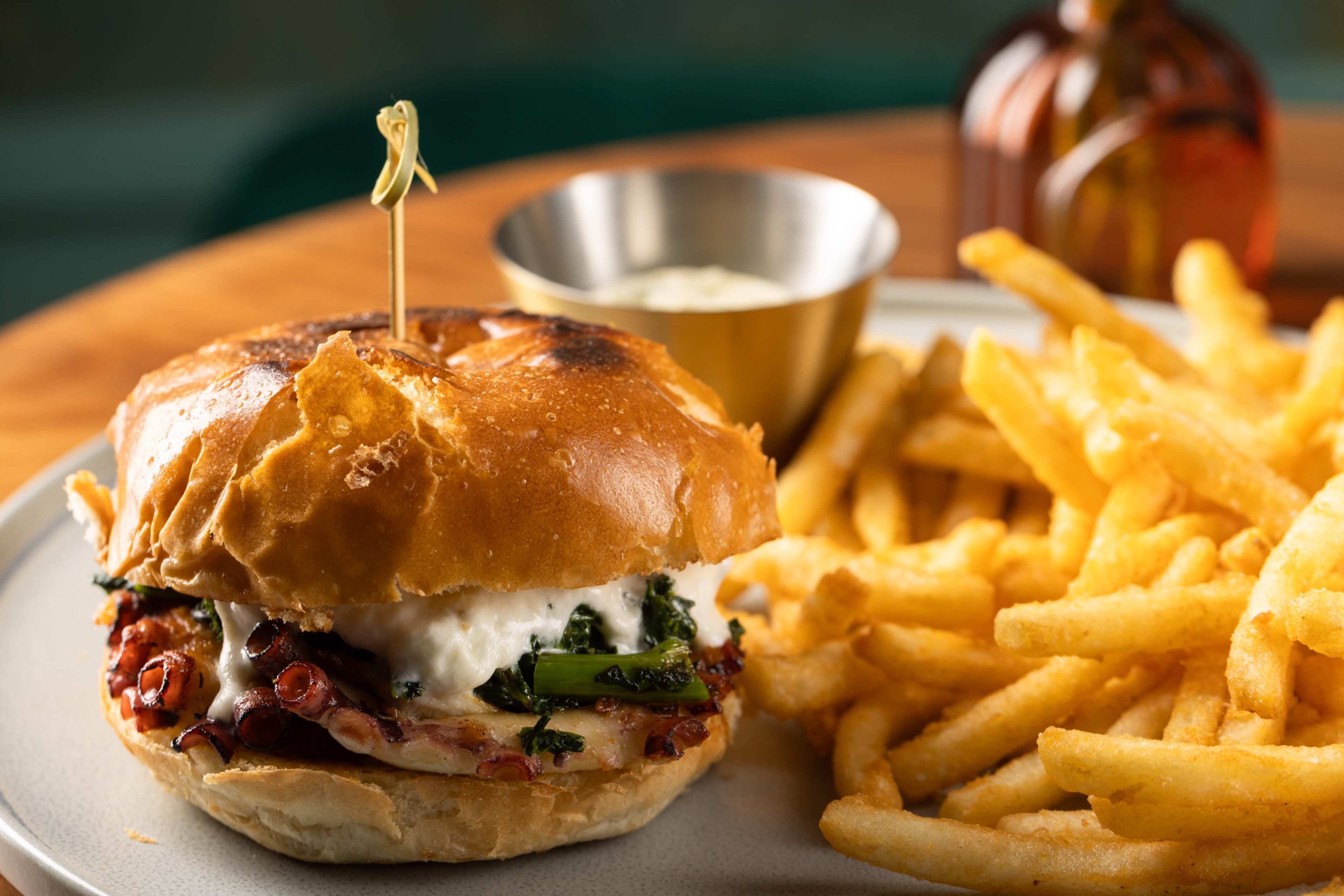 A toasted sandwich with greens, sauce, and meat is served alongside a portion of golden French fries and a small bowl of dipping sauce.