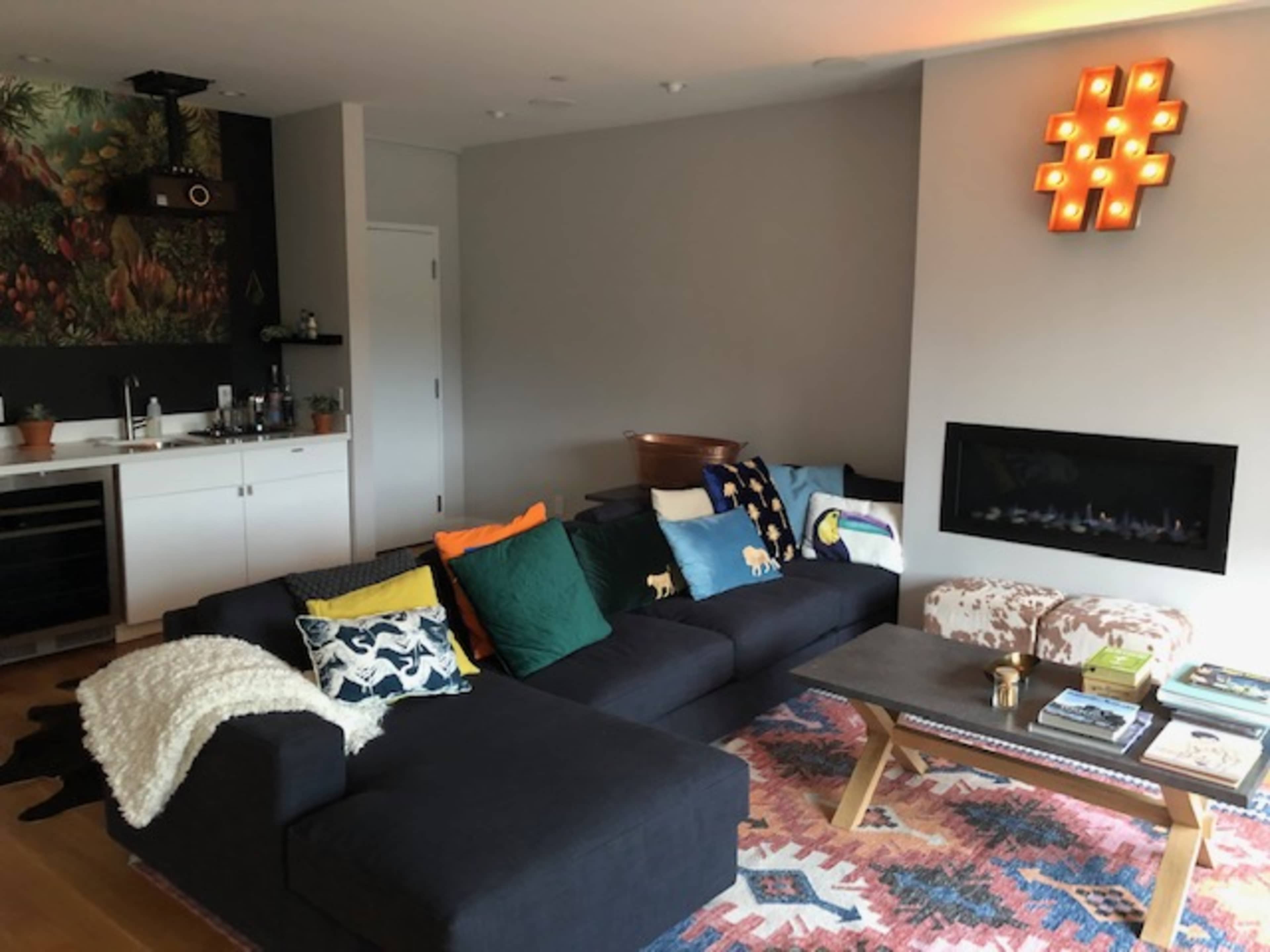 The living room features a black sectional sofa with colorful cushions, a coffee table, and a wall-mounted fireplace, set against a backdrop of patterned carpet and a mural.