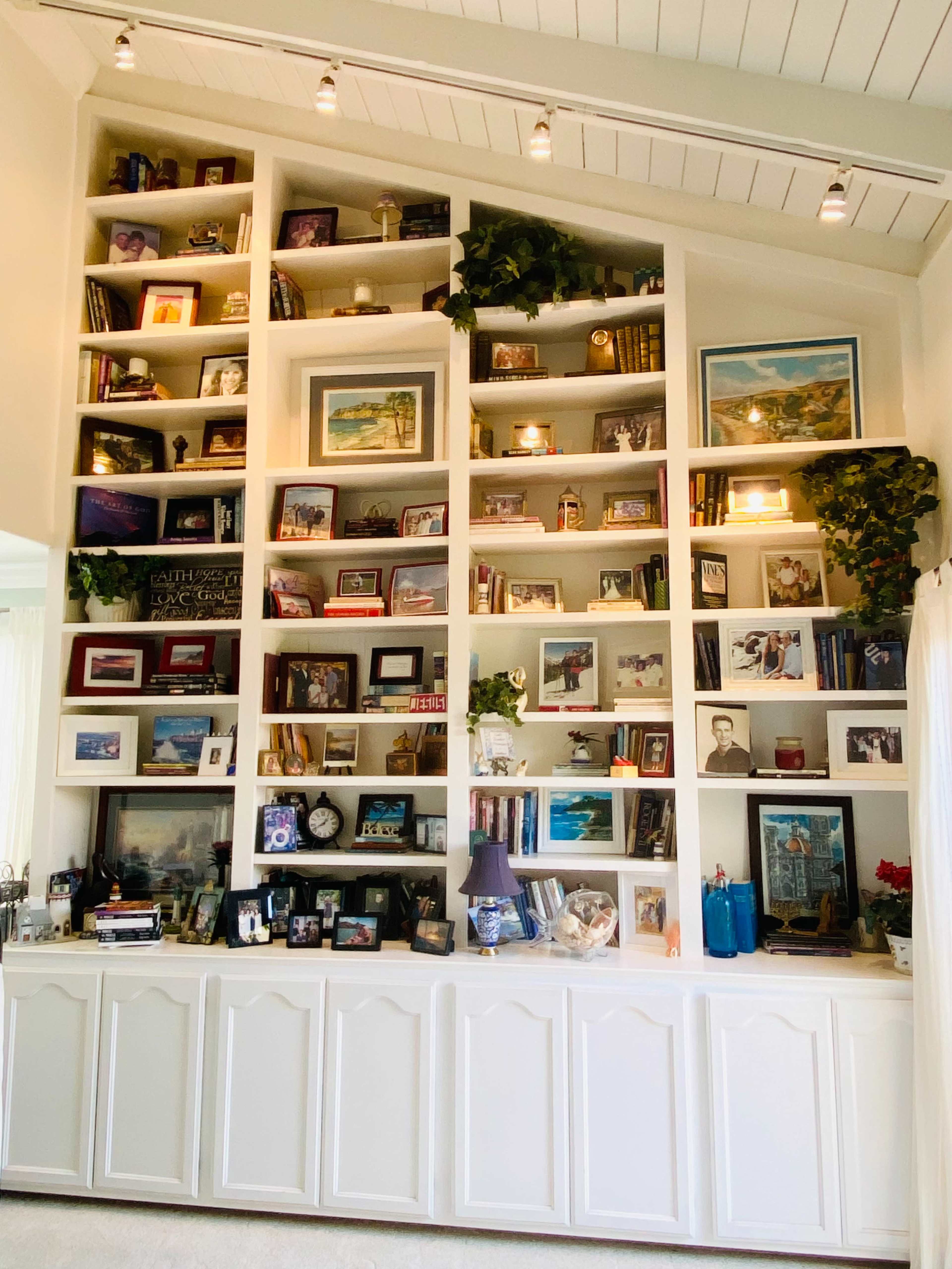 A large, well-organized bookshelf filled with books, framed photos, and decorative items is mounted on the wall of a room with light-colored walls and a high ceiling.
