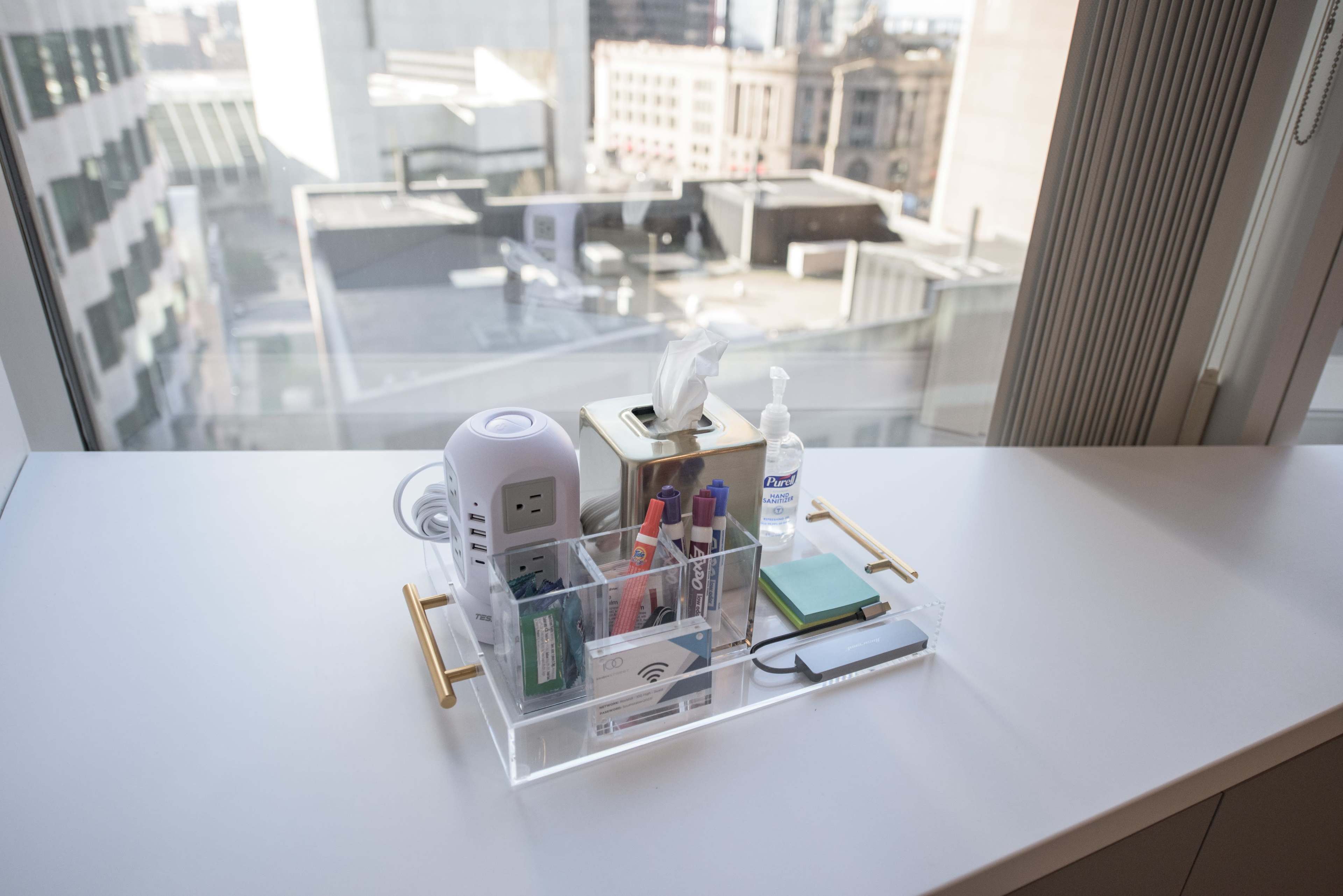 A clear acrylic tray containing various office supplies, including pens, tissue, hand sanitizer, and a small notepad, arranged on a white desk near a window with a city view.