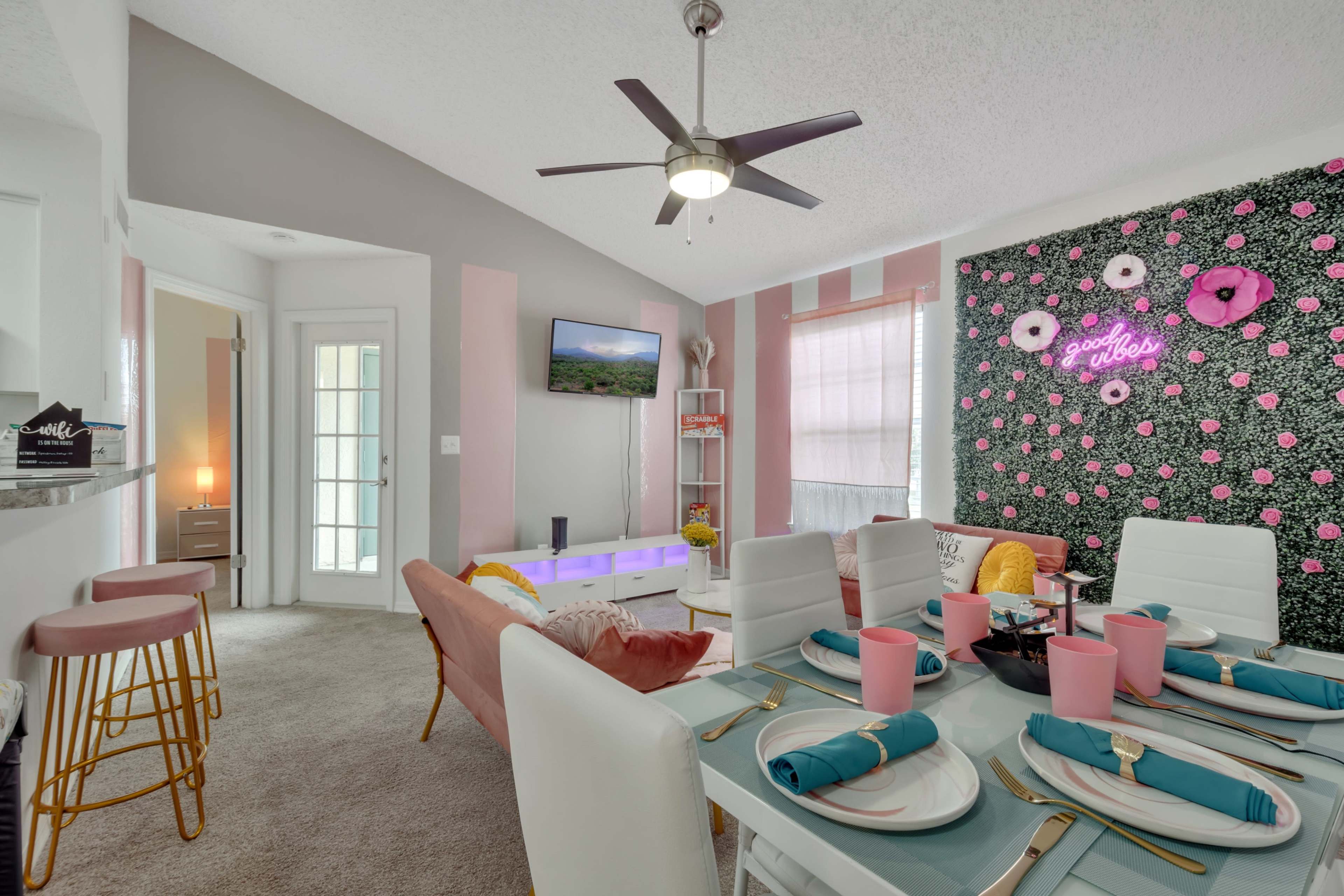 The image shows a modern dining area with a table set for a meal, featuring pink tableware and a wall adorned with floral decorations.