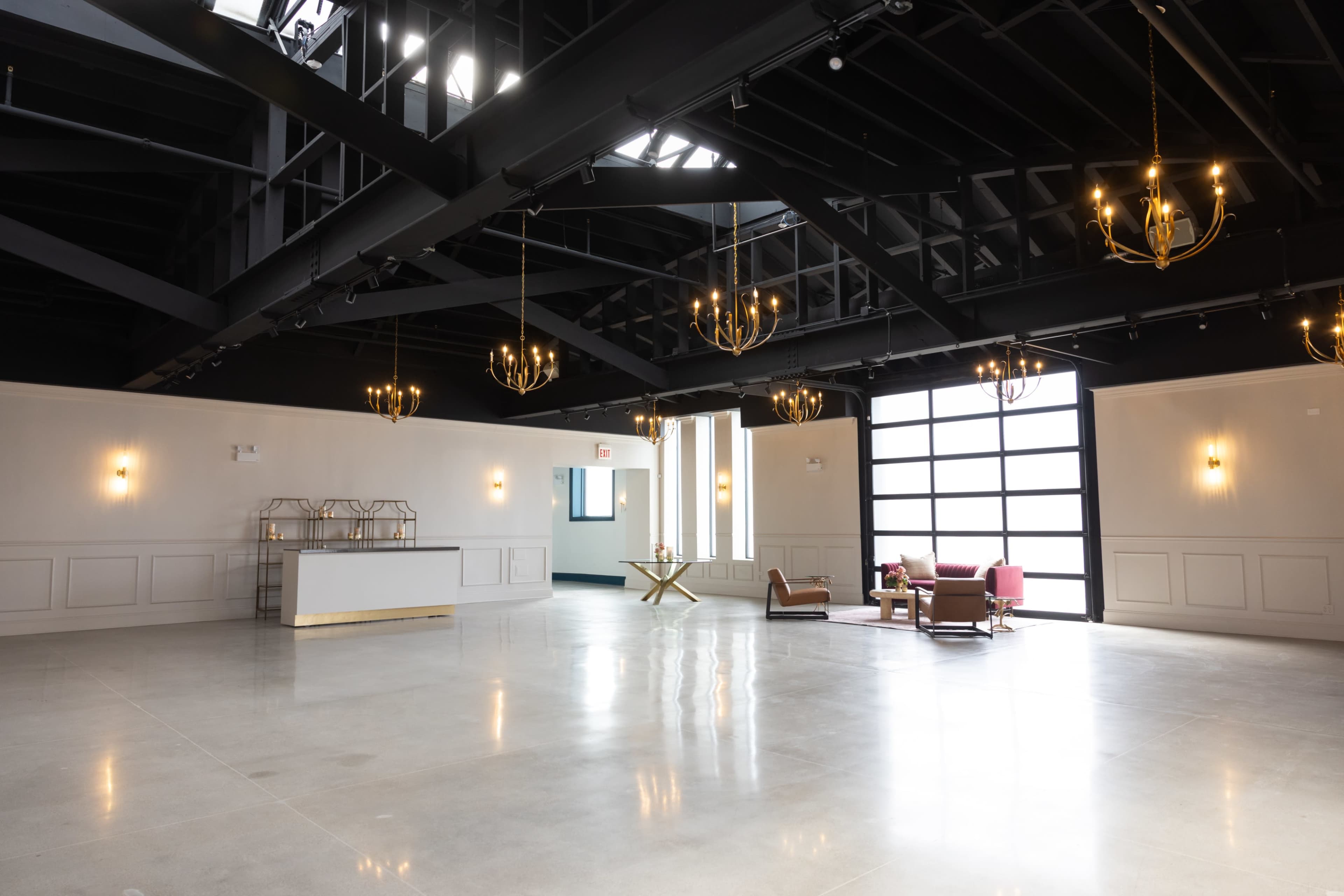 The image shows a spacious, modern interior featuring high ceilings with exposed beams, elegant lighting fixtures, and a minimalist design with seating arrangements and an event counter.