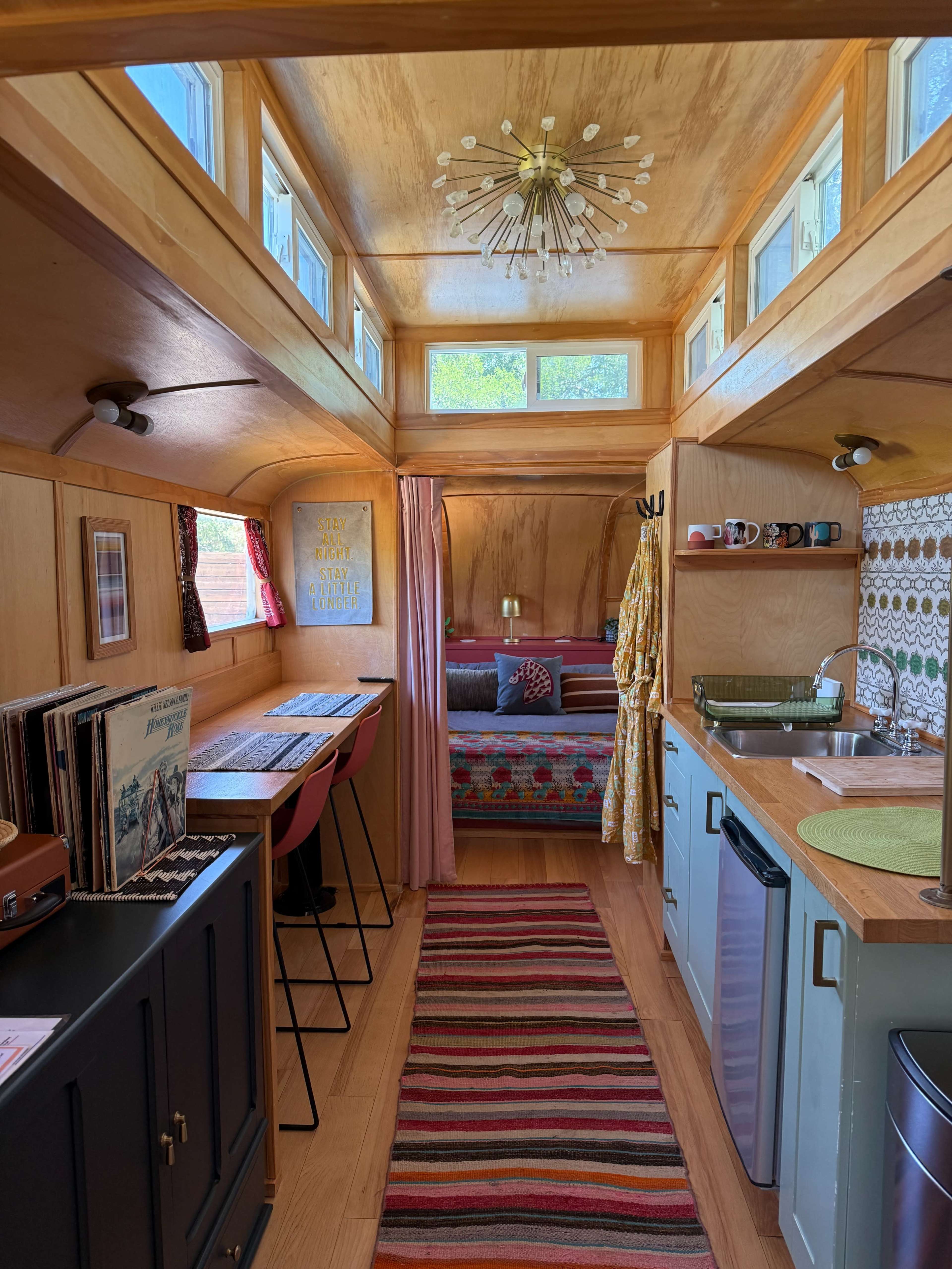 The image shows the interior of a small, stylishly designed living space featuring wooden walls, a cozy seating area, a dining table with chairs, and a compact kitchen.
