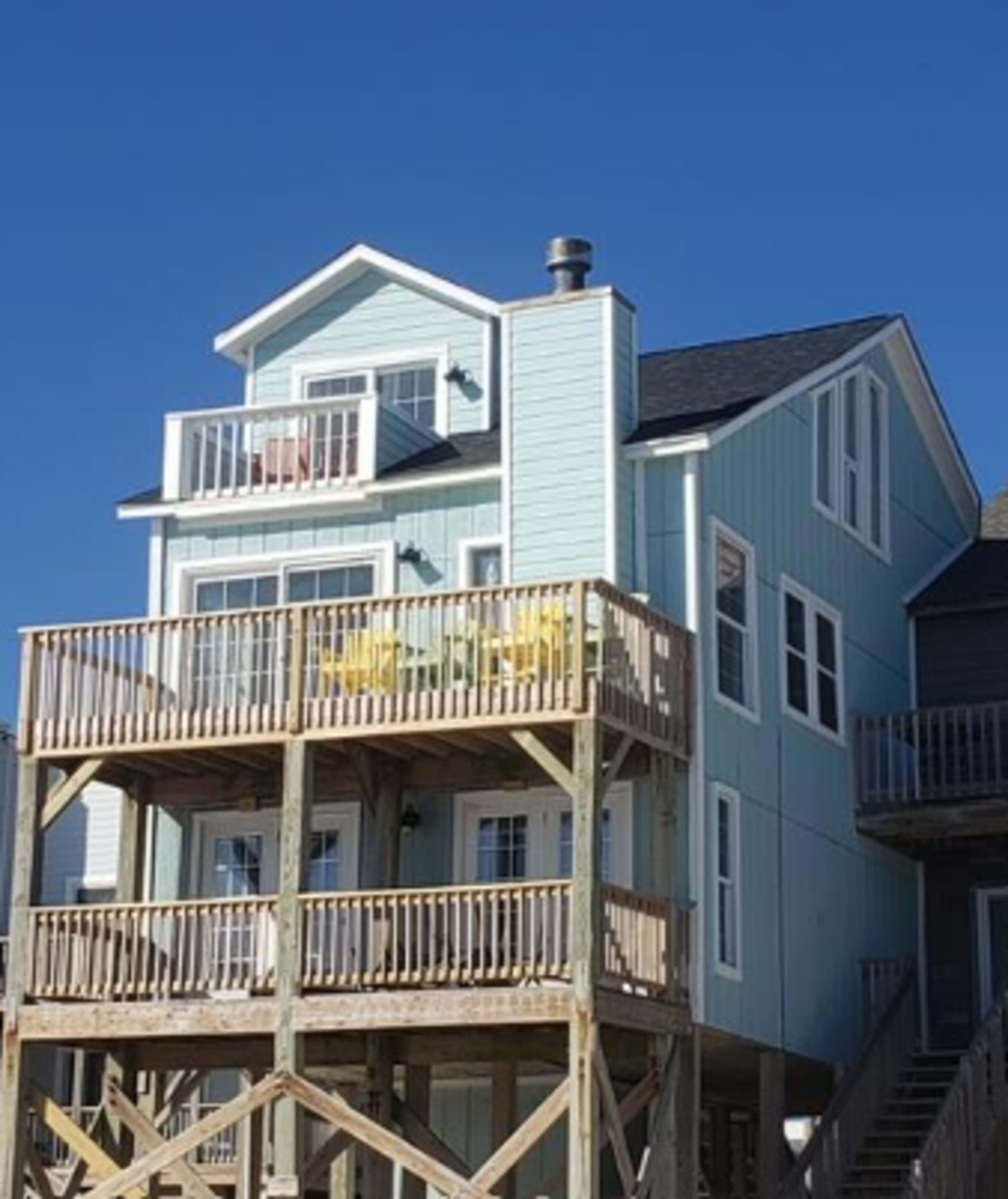 A two-story beach house with a light blue exterior and a wooden deck features two yellow chairs on the upper balcony.
