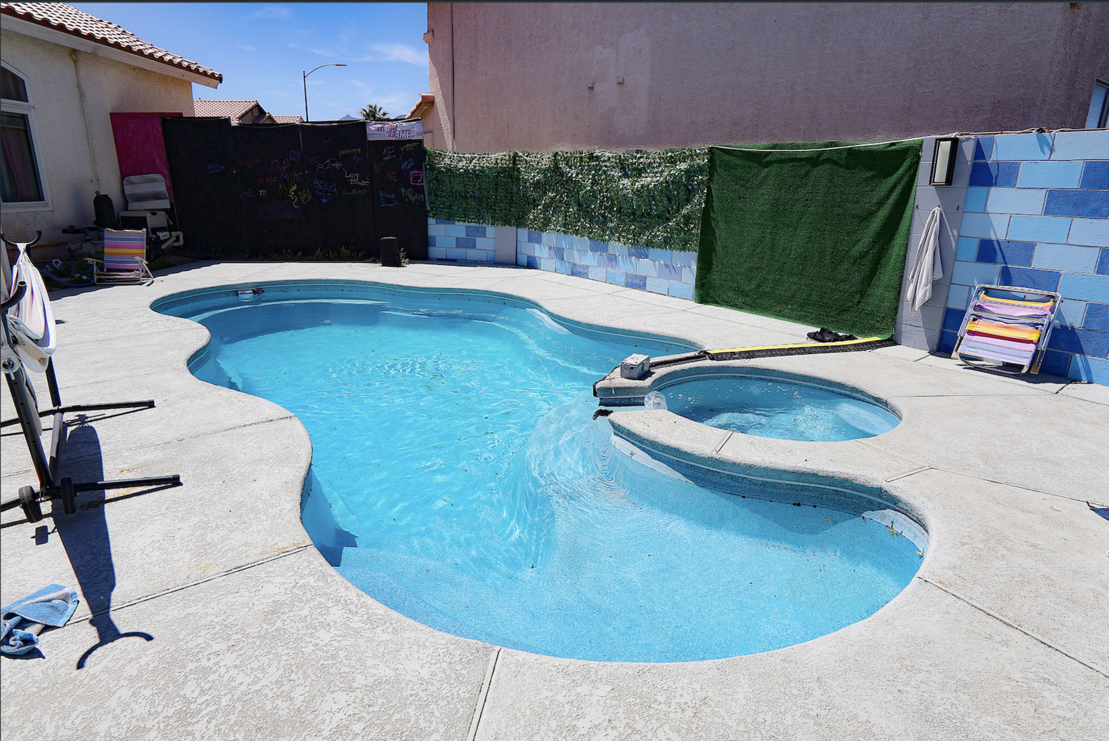 The image shows a swimming pool with a spa attached, surrounded by concrete and green privacy screens.