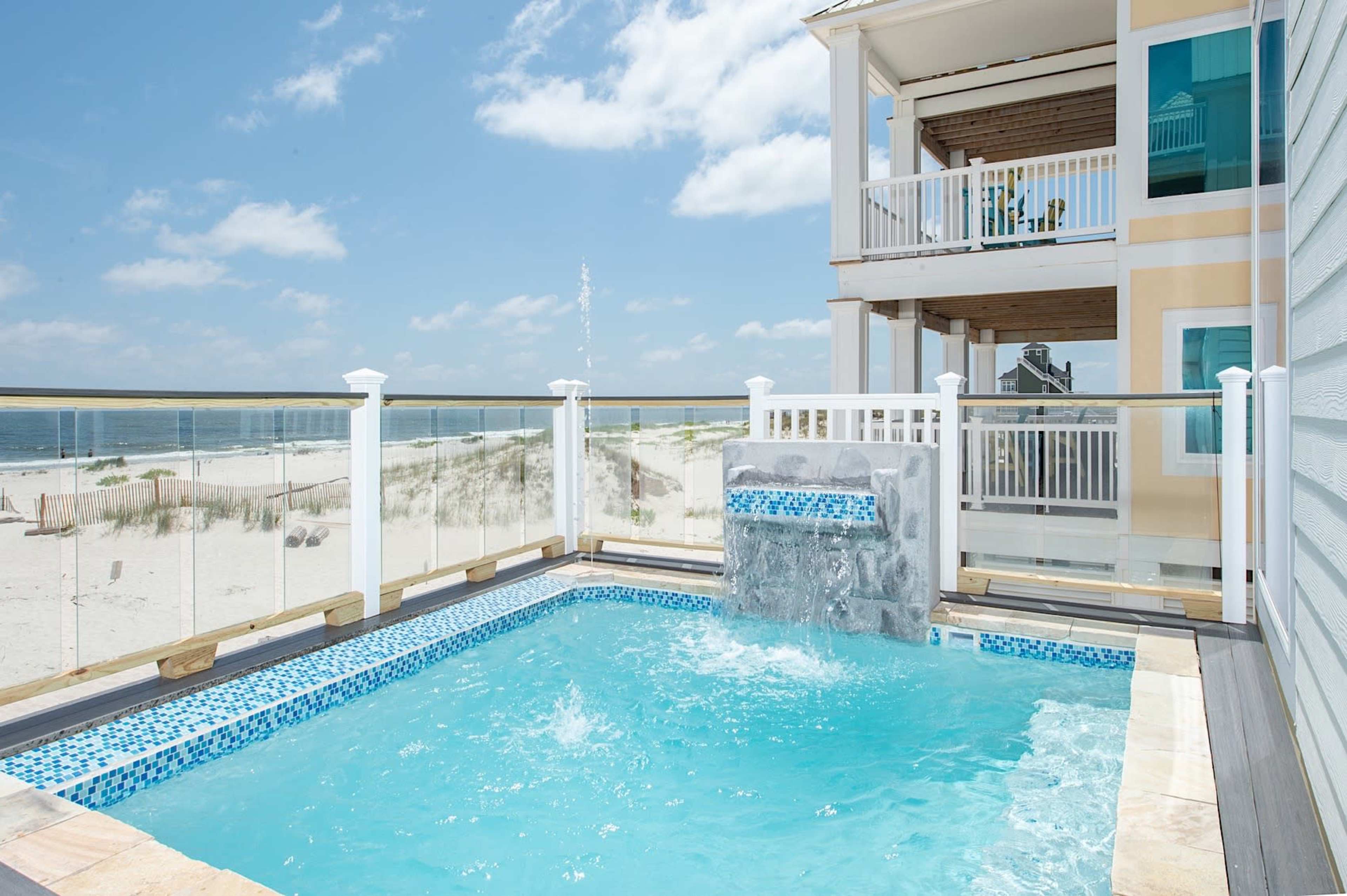 A small pool with a water feature located next to a beach house, overlooking a sandy beach and ocean.