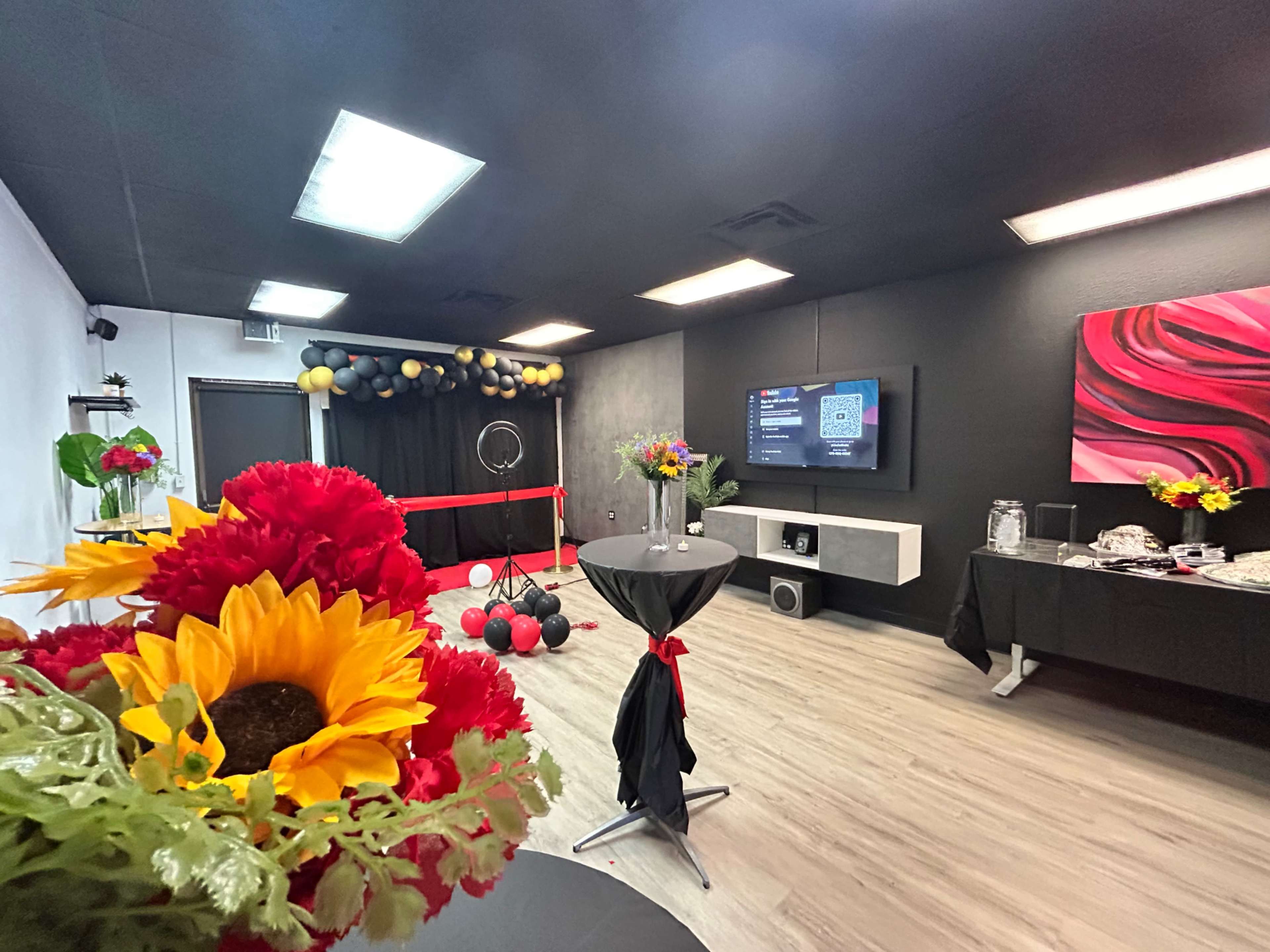 A decorated event space features tables with floral arrangements, a large screen on the wall, and a backdrop of balloons.