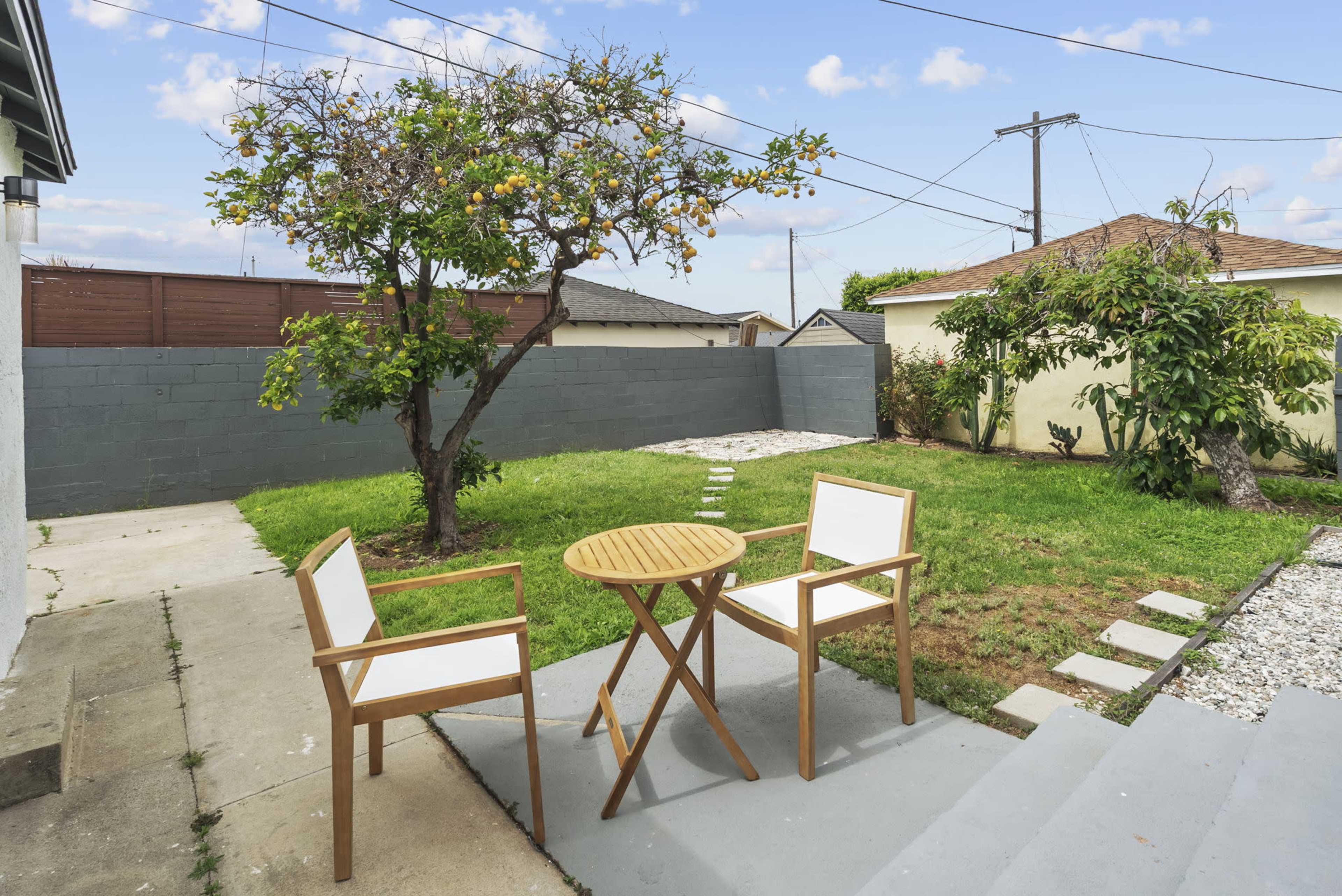The image shows a backyard with a small wooden table and two chairs, a fruit tree, and a grassy area surrounded by concrete and stone paths.