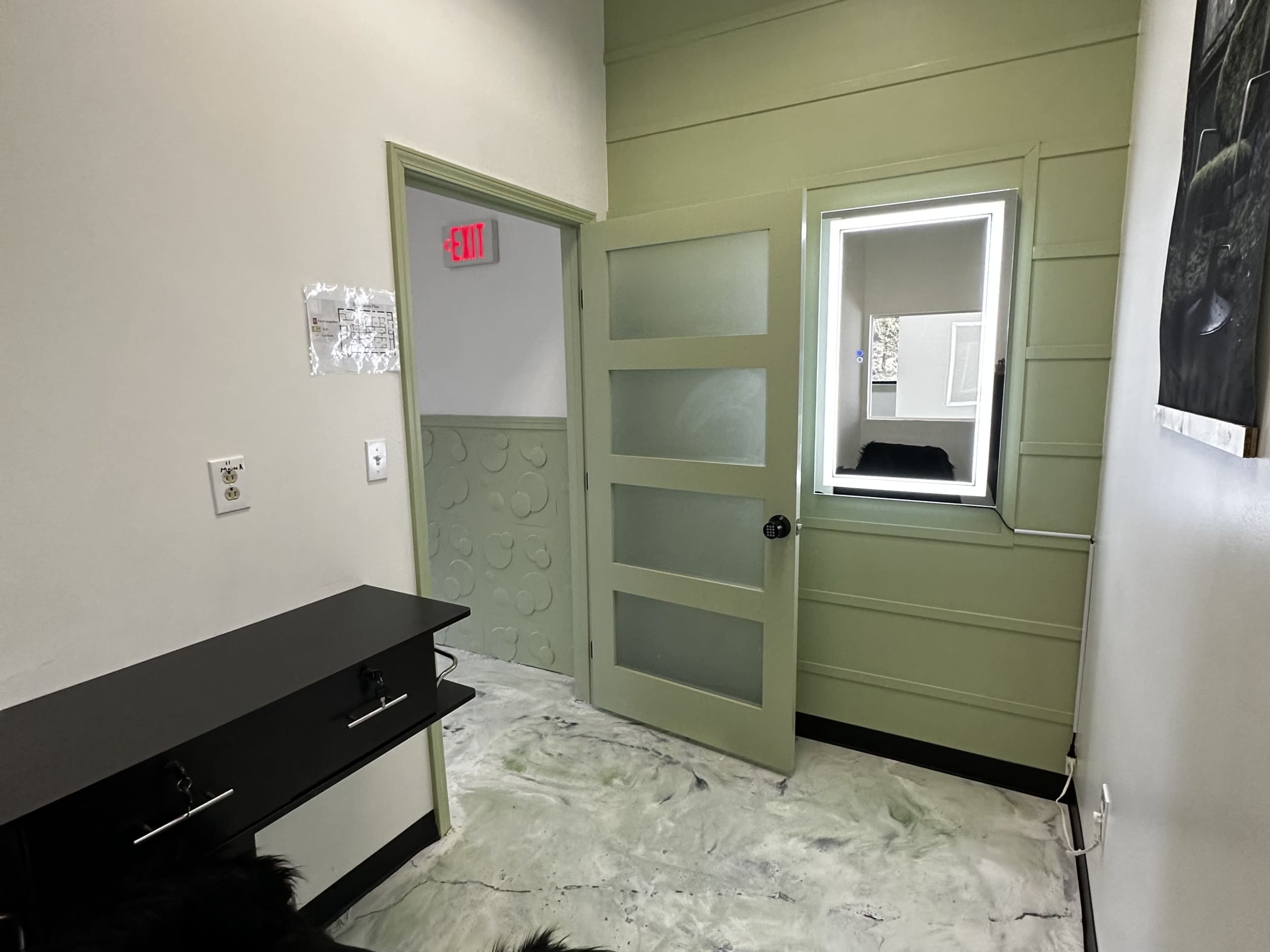 The image shows a small, well-lit hallway with a partially open green door leading to a room, a window on the right, and a dark cabinet on the left.