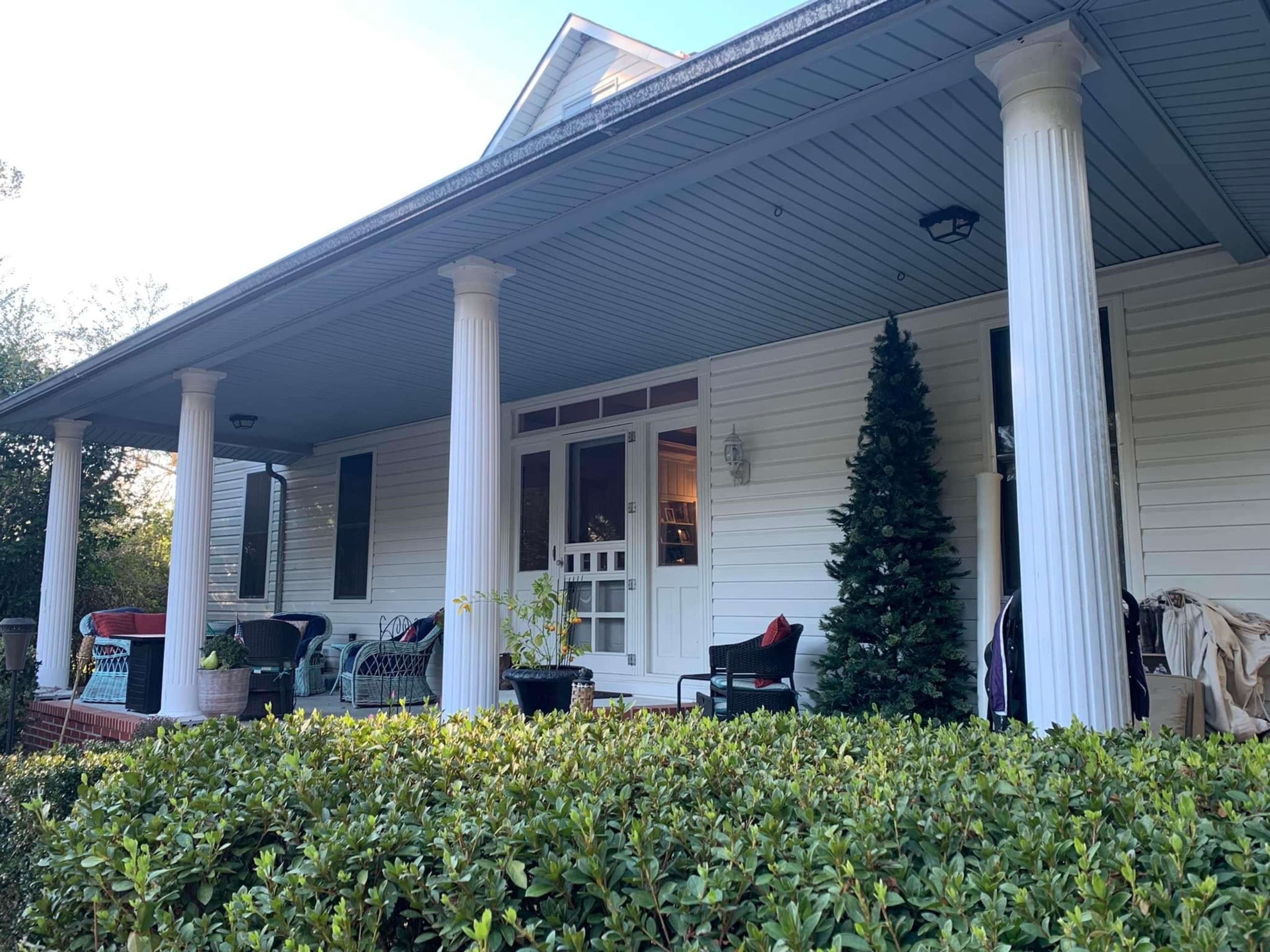 The image shows a house with a front porch supported by white columns and a green hedge in the foreground.
