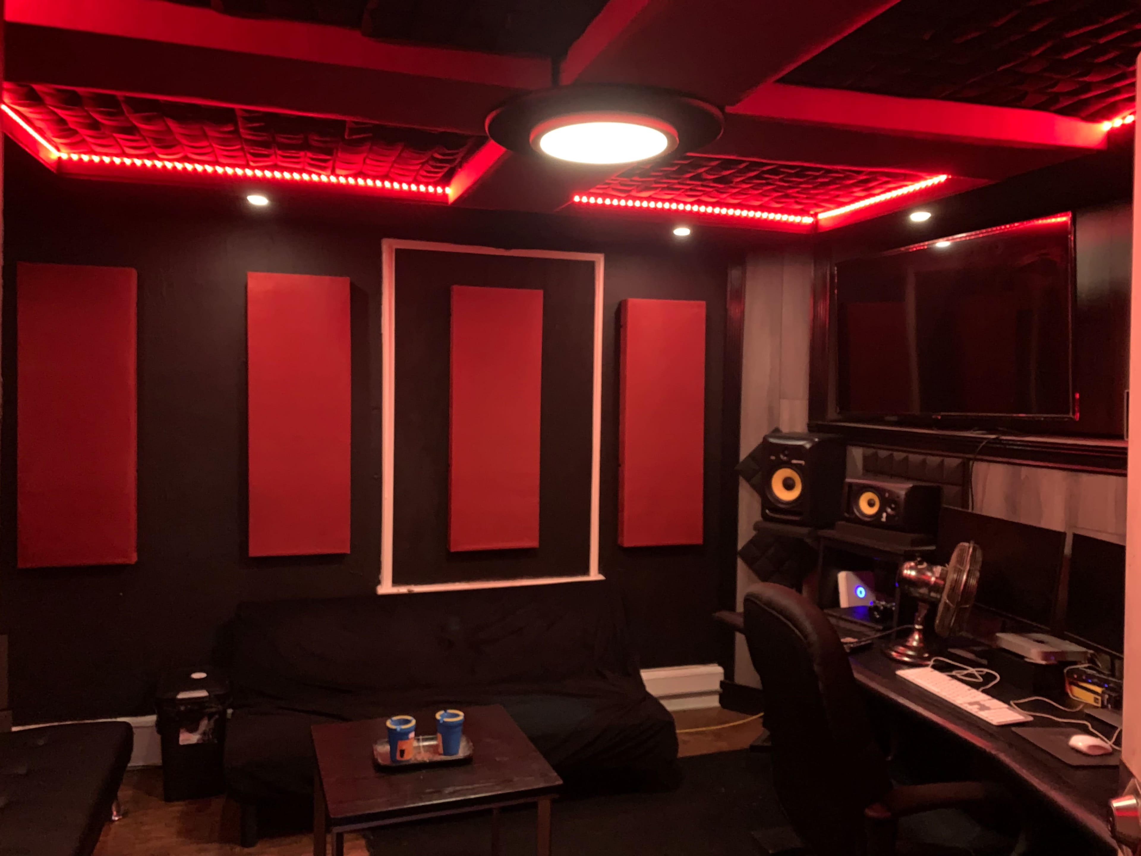 A recording studio with black walls, red soundproofing panels, a couch, and a desk equipped with audio equipment and speakers.