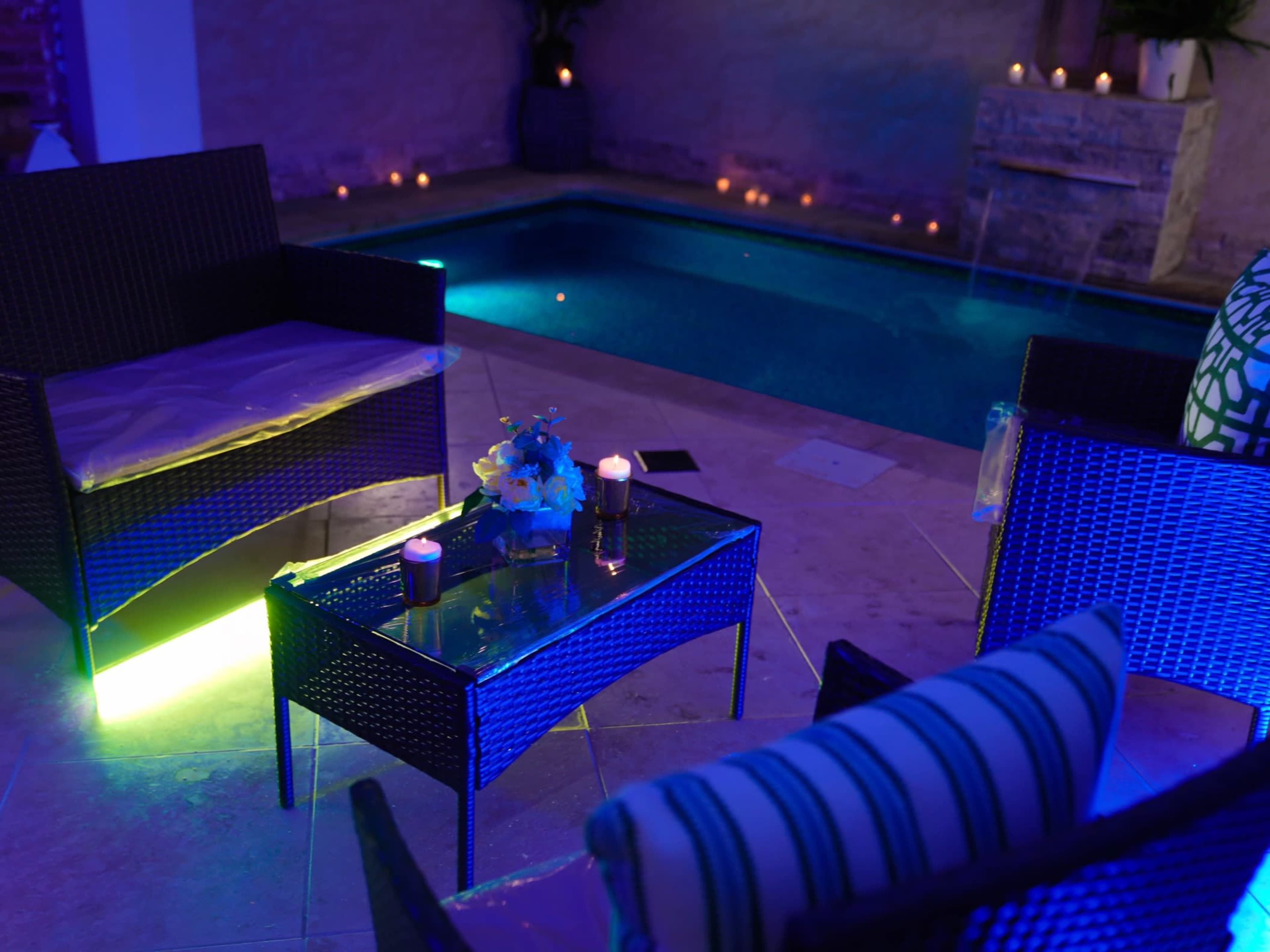 A serene poolside setting featuring two wicker chairs and a glass-top table adorned with candles and flowers, illuminated by blue lighting.