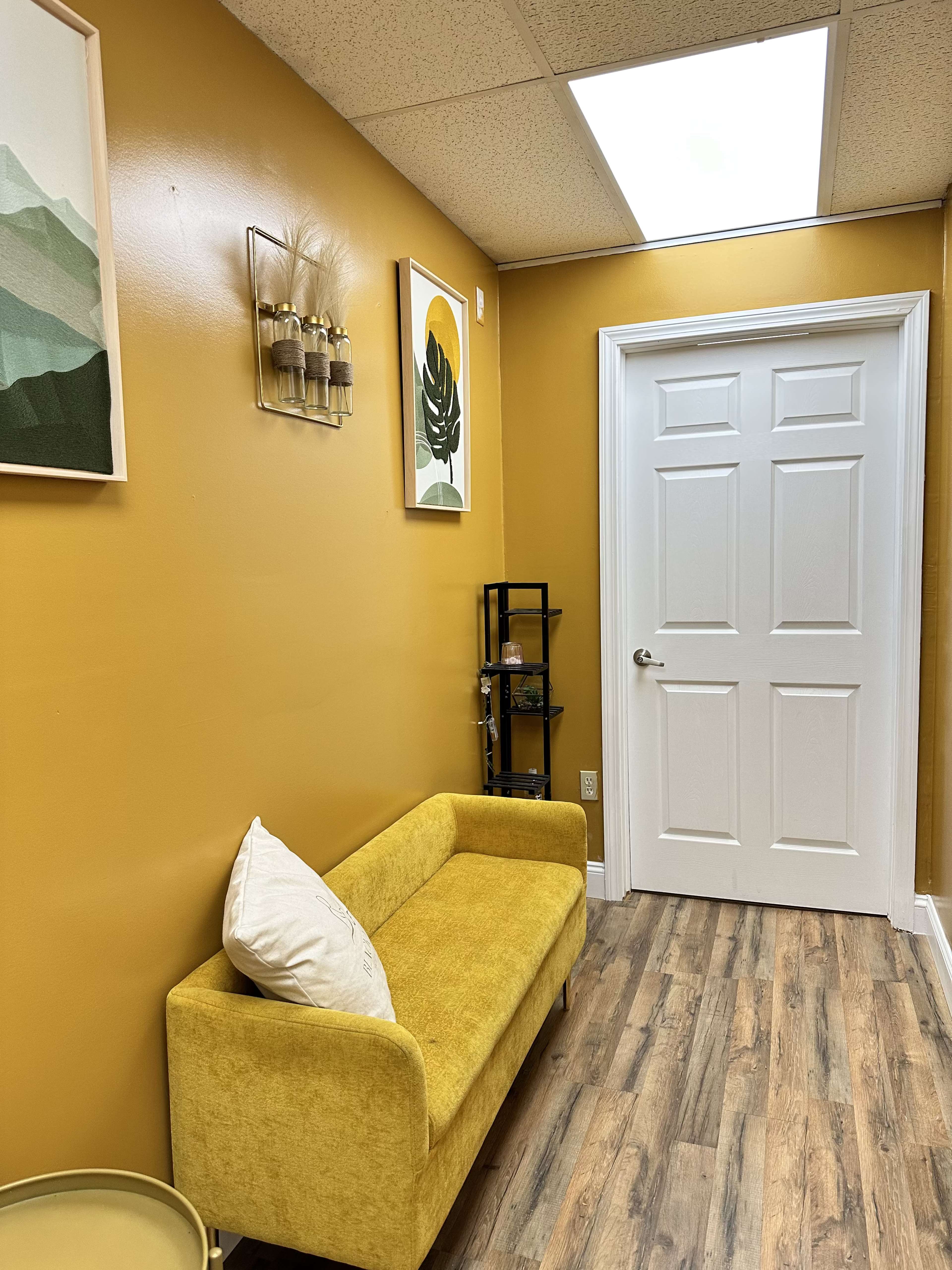 The image shows a cozy hallway with a yellow wall, a small yellow couch, and artwork on the walls, leading to a white door.