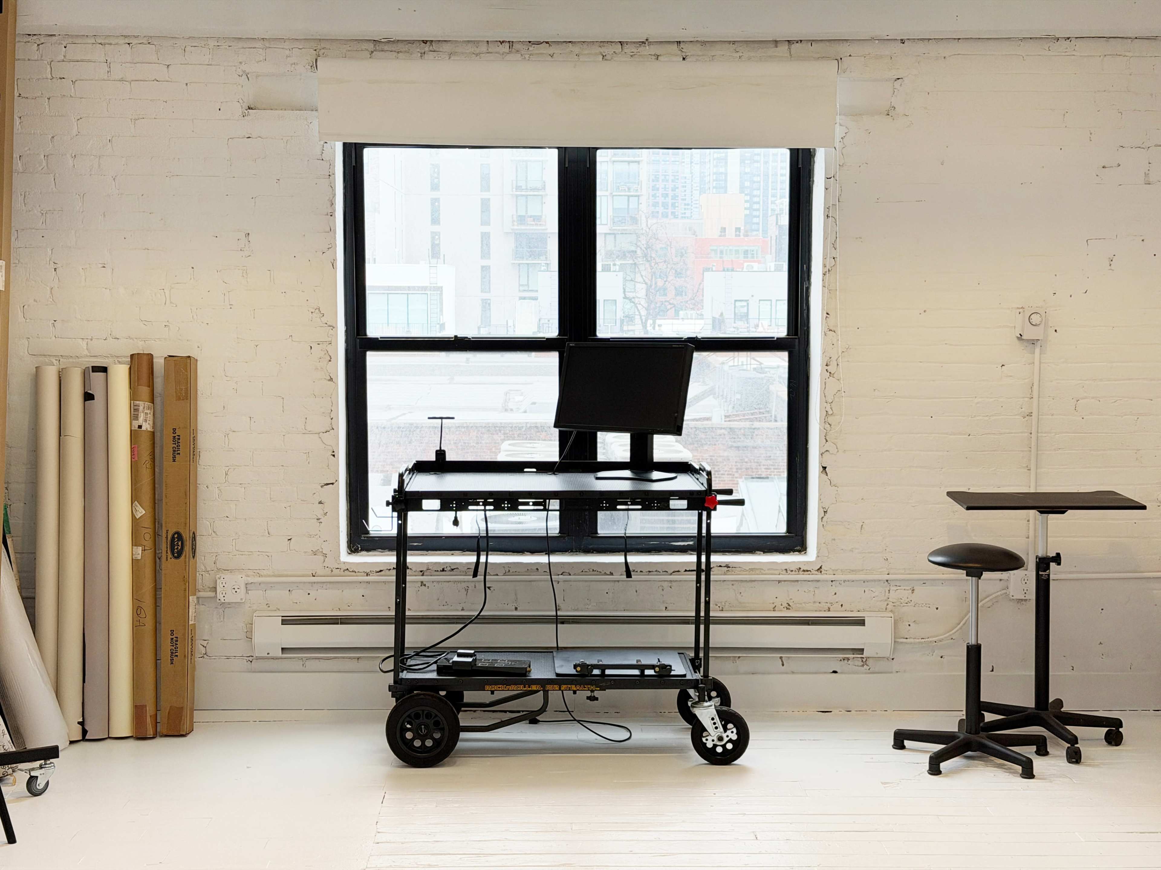 A black adjustable-height desk with a computer monitor is positioned in front of a large window in a spacious, minimalist room with white brick walls.