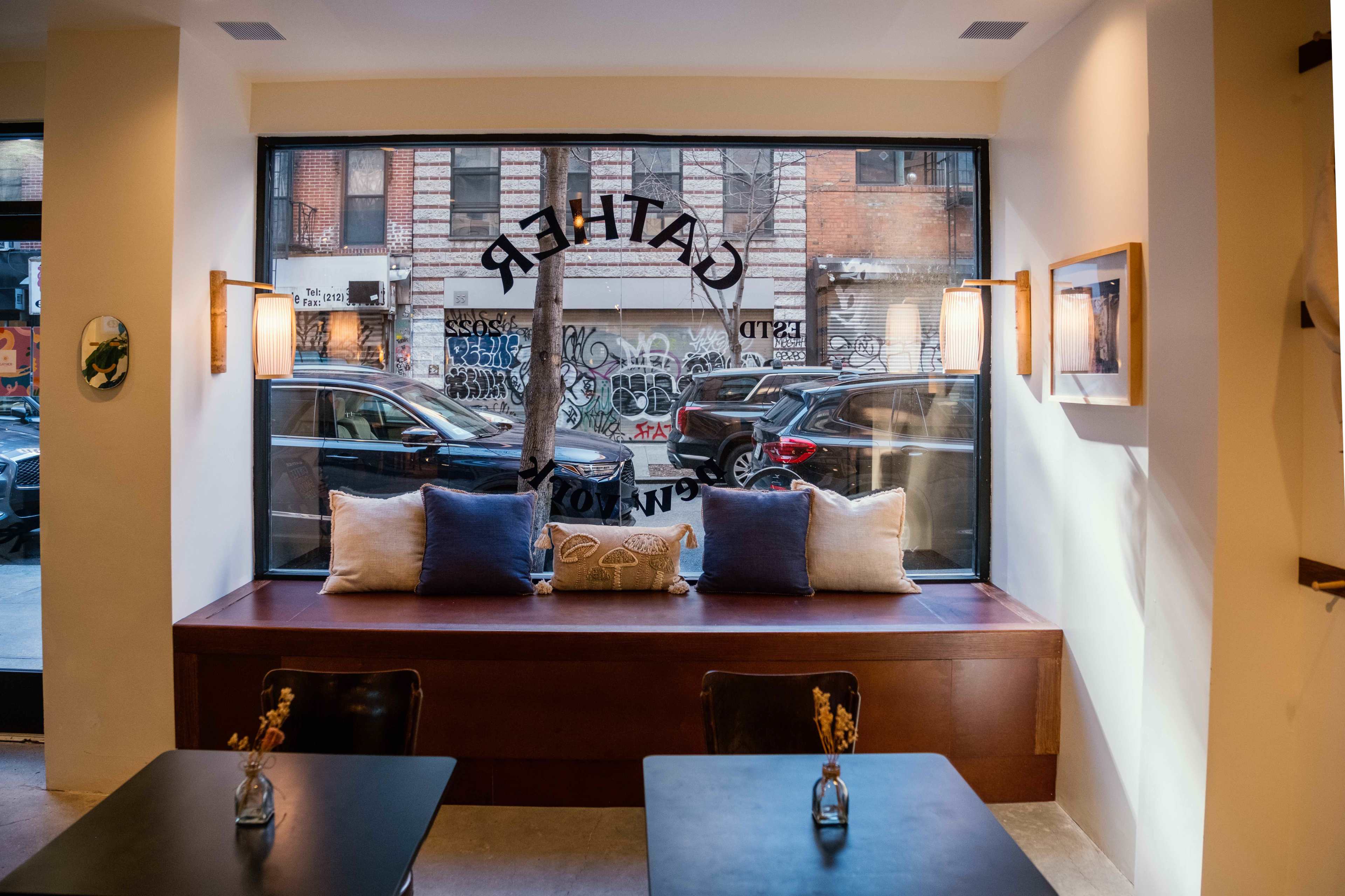 A cafe features a window seat with decorative pillows and a view of graffiti-covered buildings and parked cars outside.