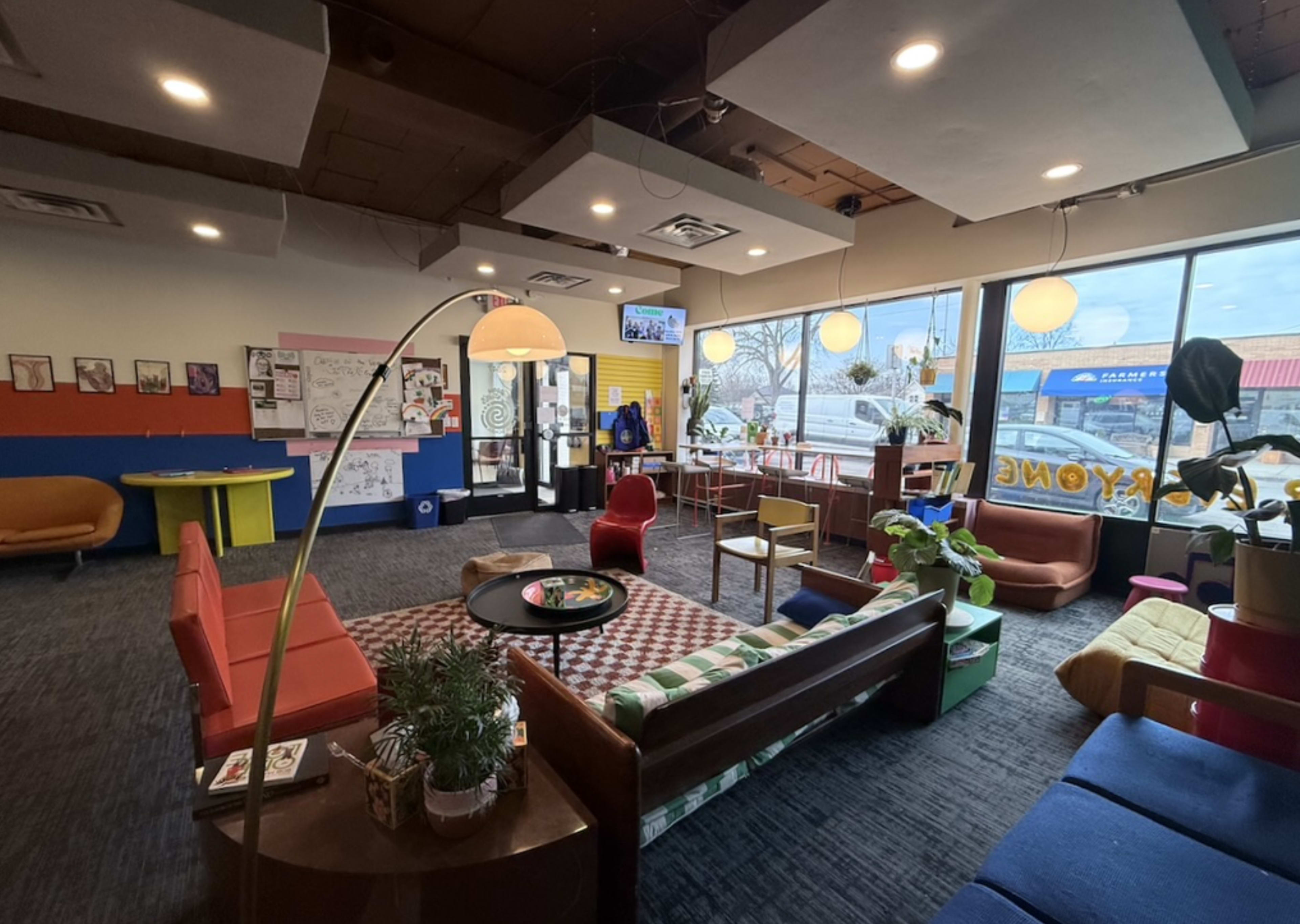 The image shows a brightly lit café interior with a variety of seating arrangements, including colorful sofas and chairs, and a large window displaying the outdoor view.