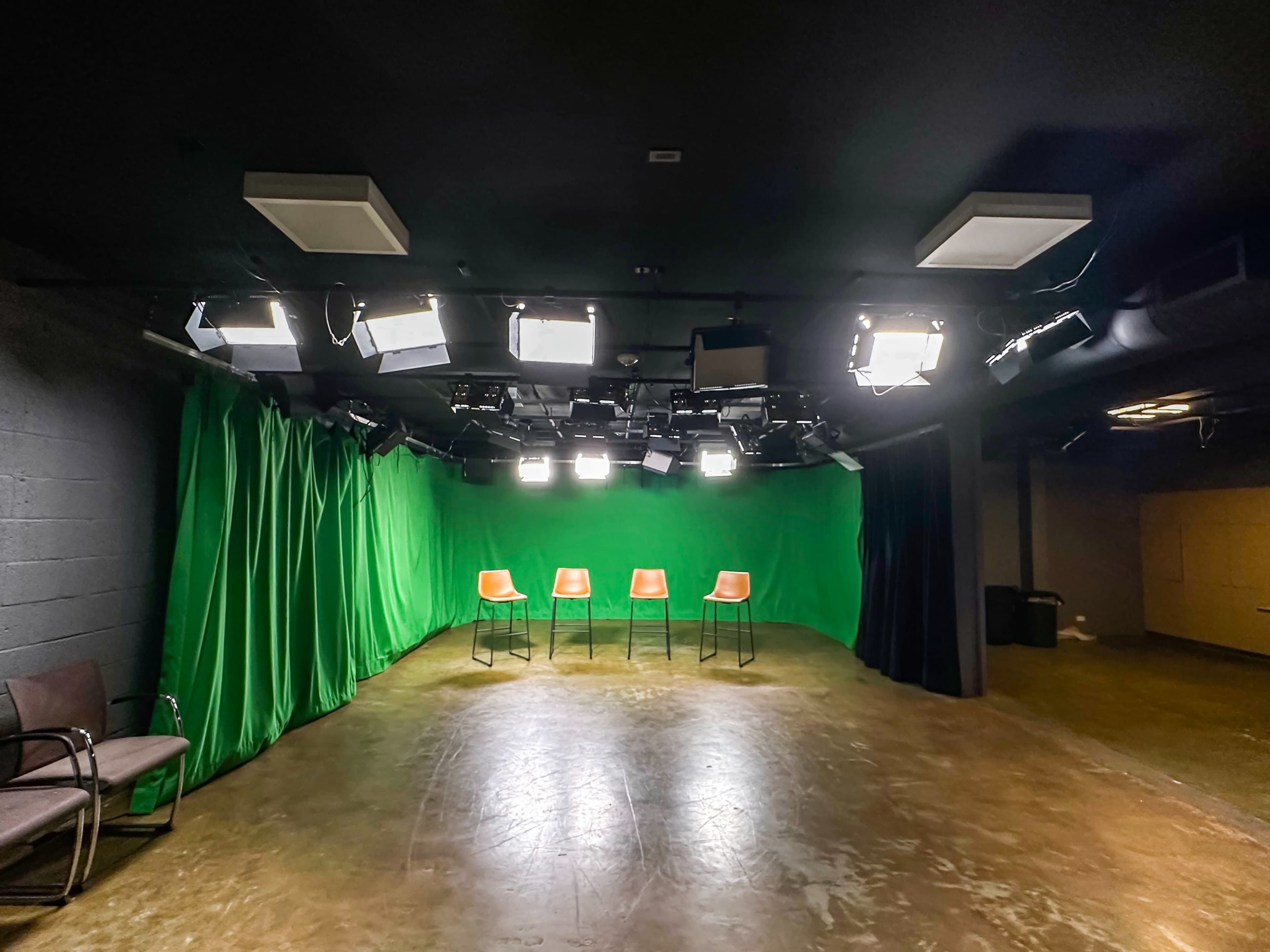 A studio space with four brown chairs arranged in front of a green backdrop, illuminated by overhead lights.