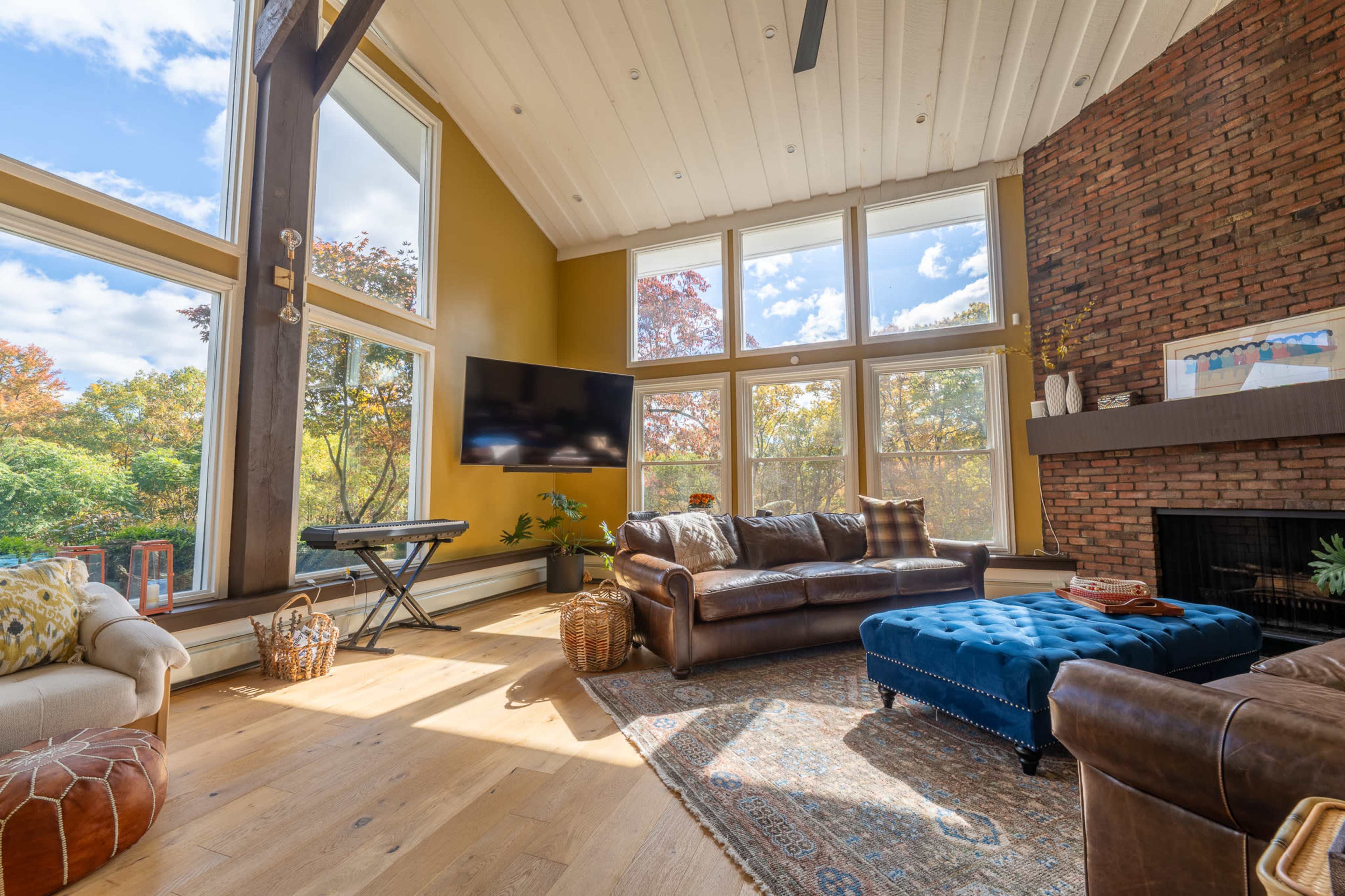 A spacious living room features large windows, a dark leather sofa, a blue ottoman, and a brick fireplace surrounded by autumn-colored foliage outside.
