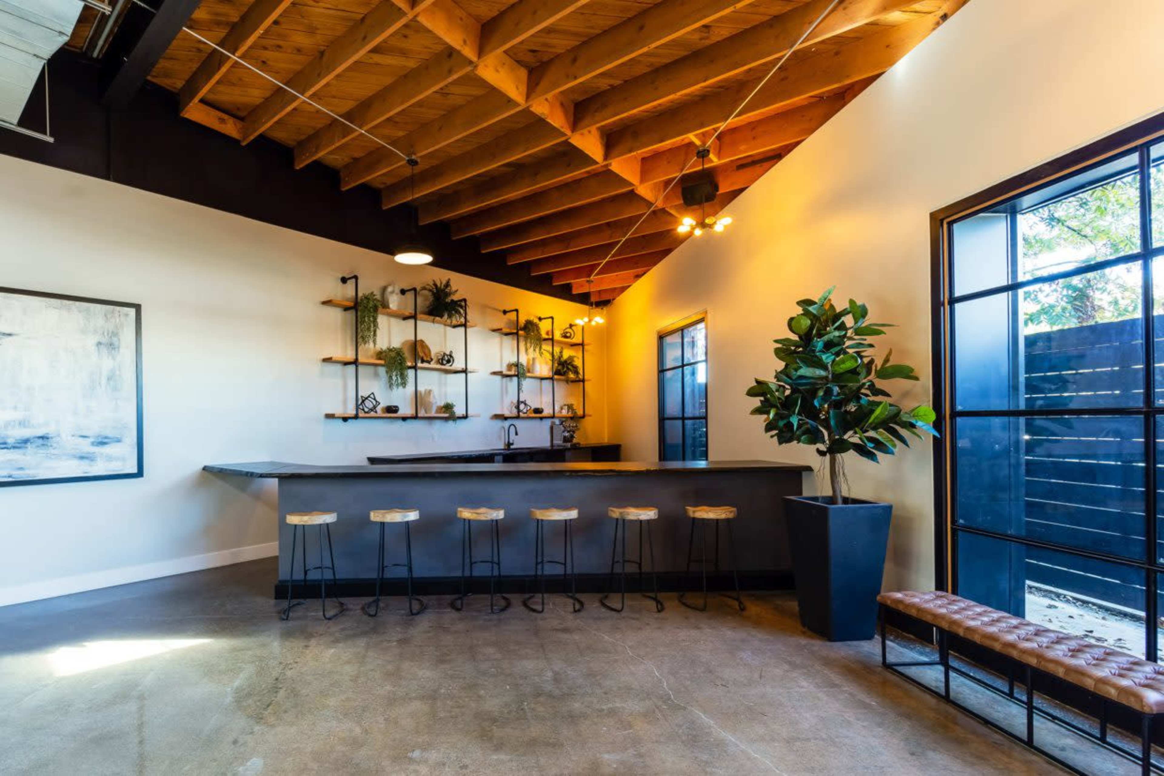 The image features a modern interior space with a bar counter, high stools, and shelves adorned with plants, set against a backdrop of wooden beams and large windows.