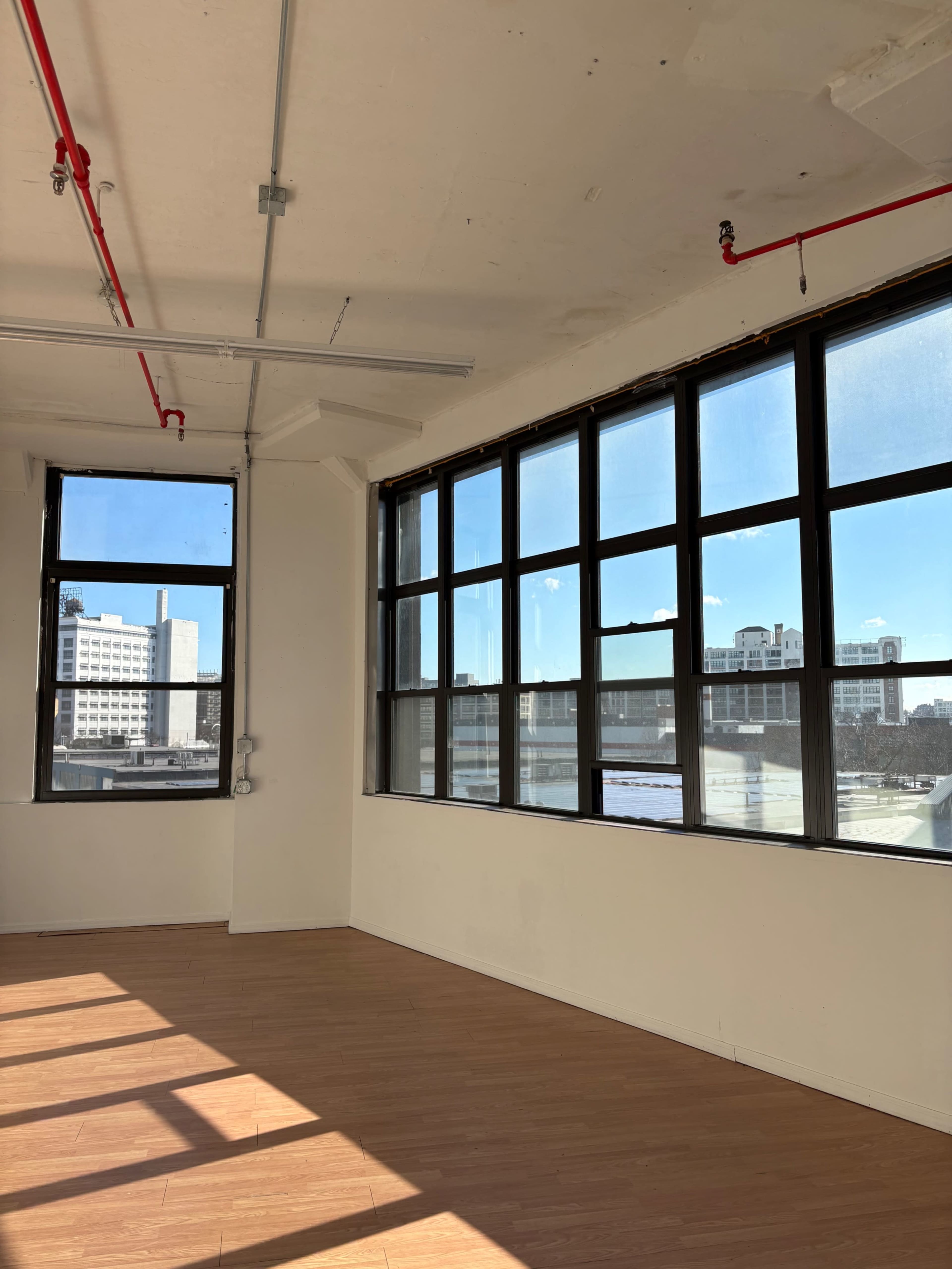 The image shows an empty room with large windows and wooden flooring, illuminated by natural light.