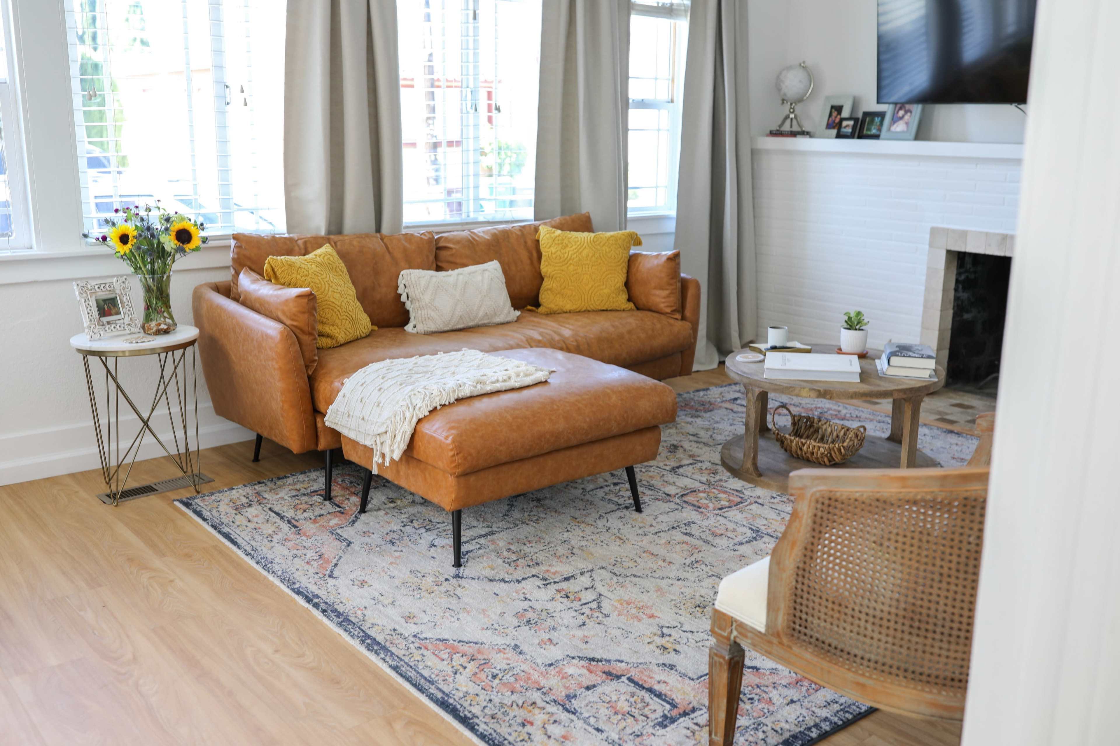 A modern living room features a brown leather sectional sofa with yellow pillows, a circular coffee table, a patterned area rug, and large windows with sheer curtains.